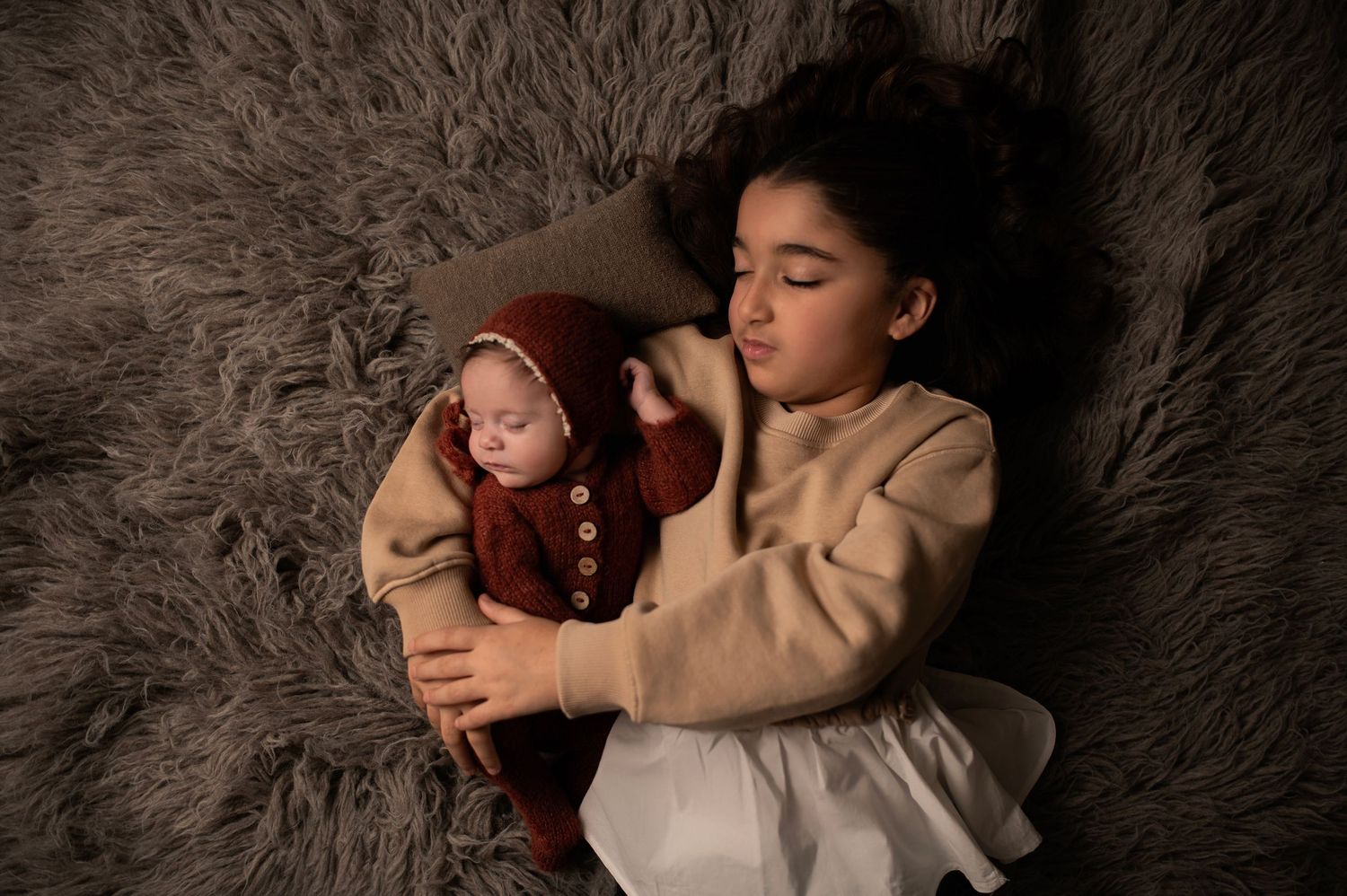 A young girl lies on a gray fur rug cradling a sleeping newborn baby dressed in a rust-colored knit outfit and bonnet.