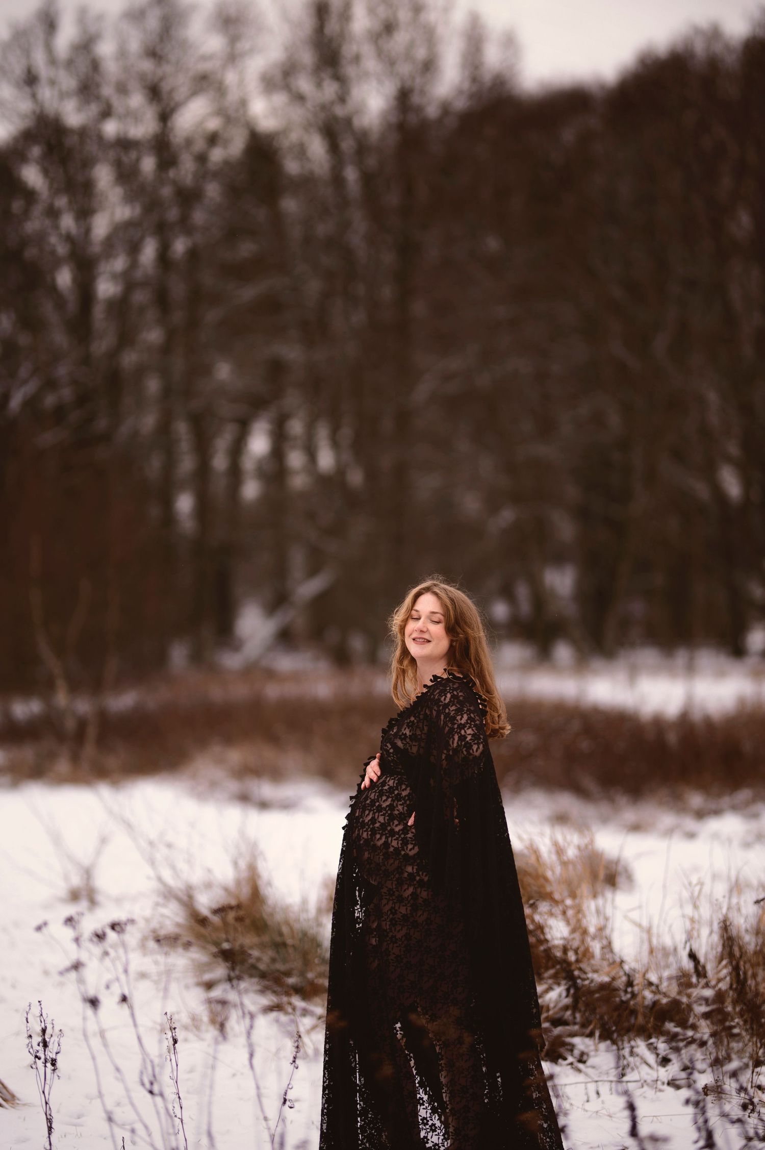 Gravidfotografering i Skåne med gravid kvinna i svart spetsklänning i snöig vinterskog.