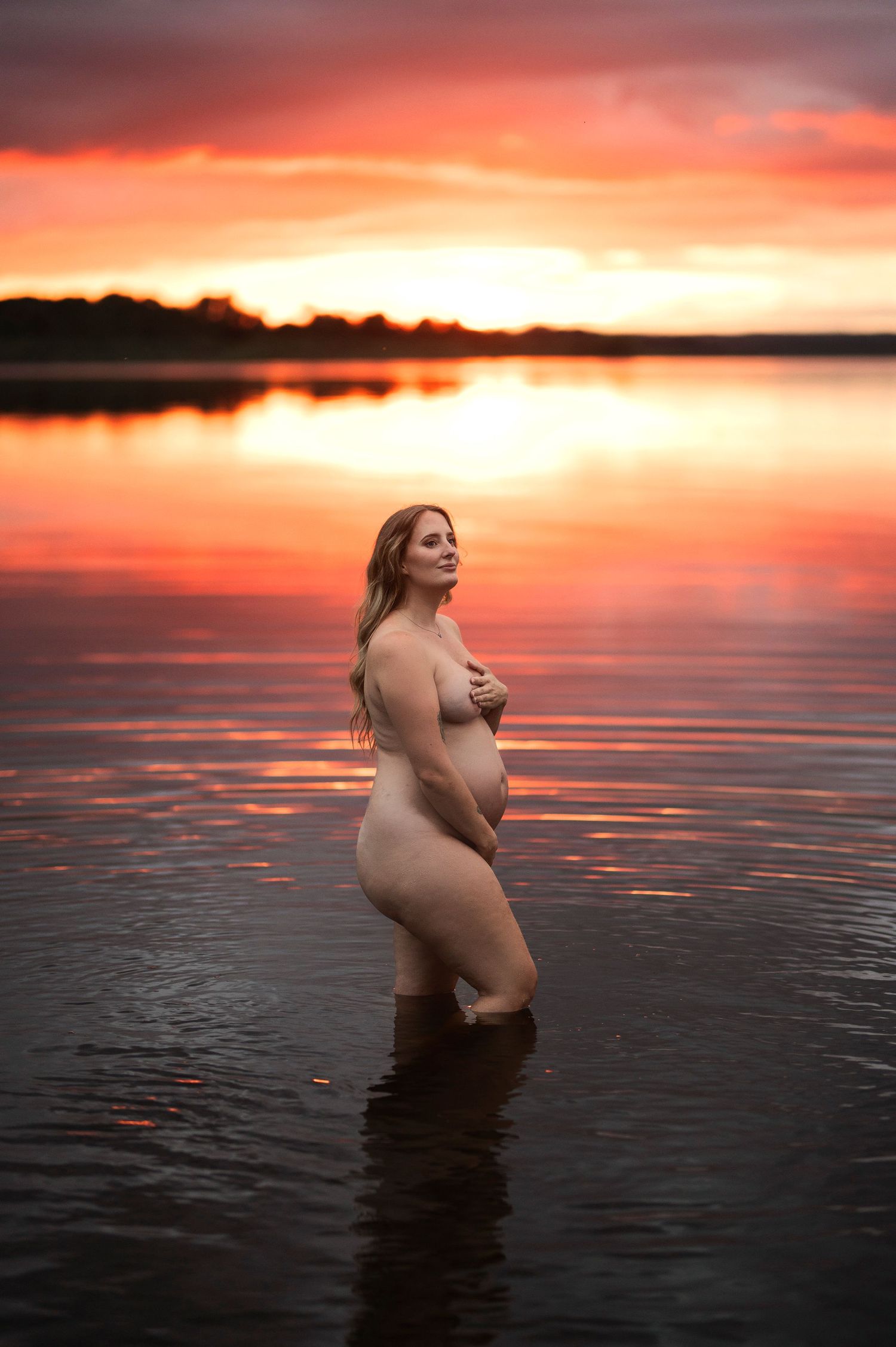 Gravidfotografering i Skåne med siluett i stilla sjövatten vid solnedgång.