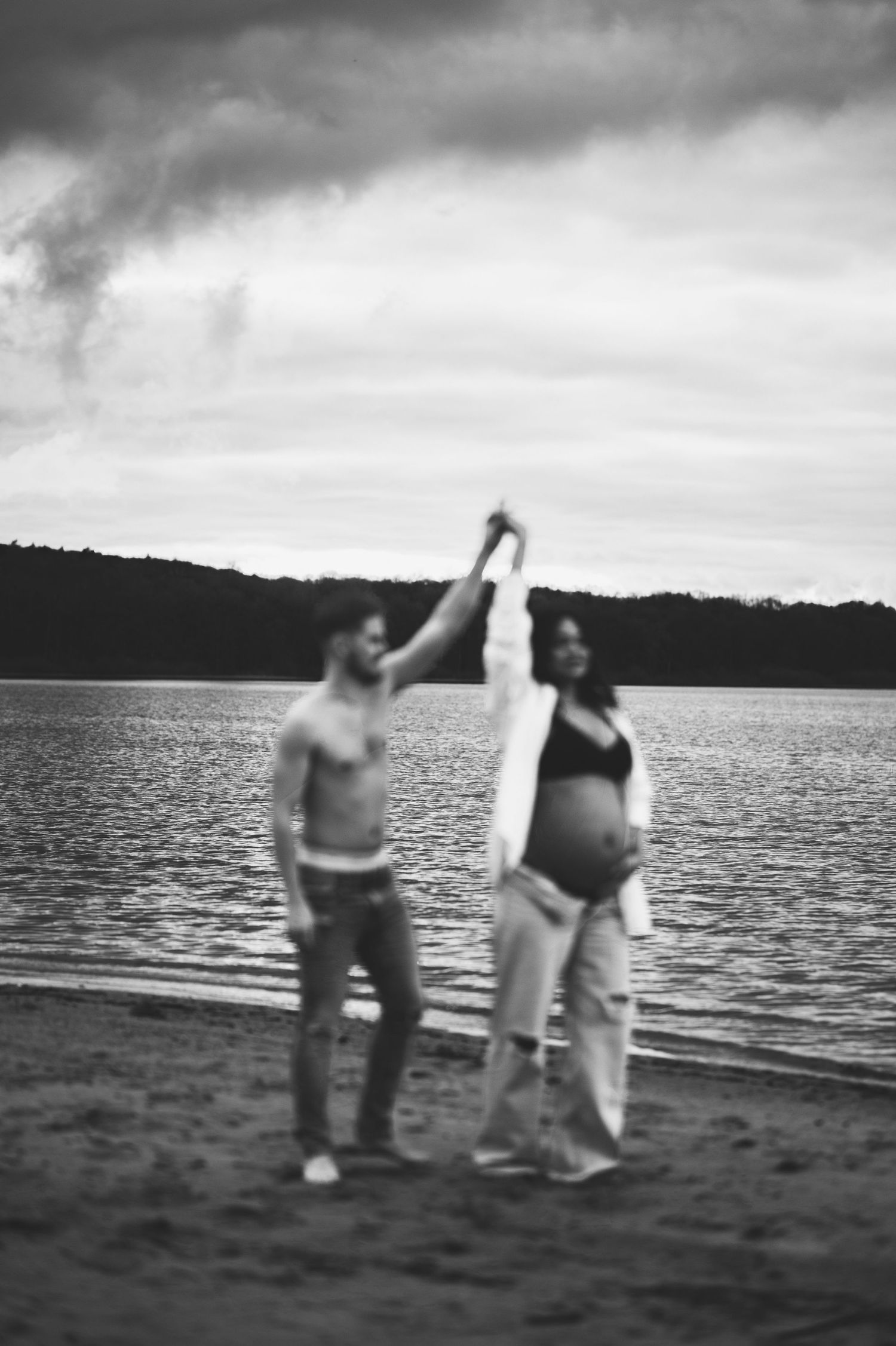 Black and white photo of two figures dancing on a beach with lake and cloudy sky in background.