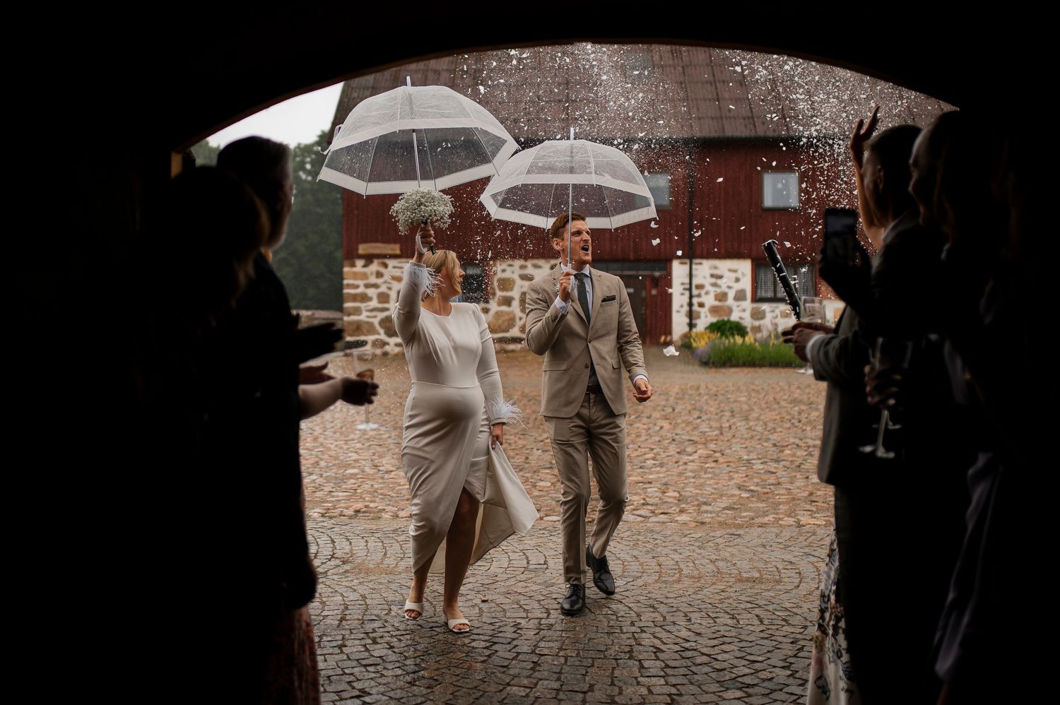 A wedding couple walks under umbrellas through a stone archway in the rain.