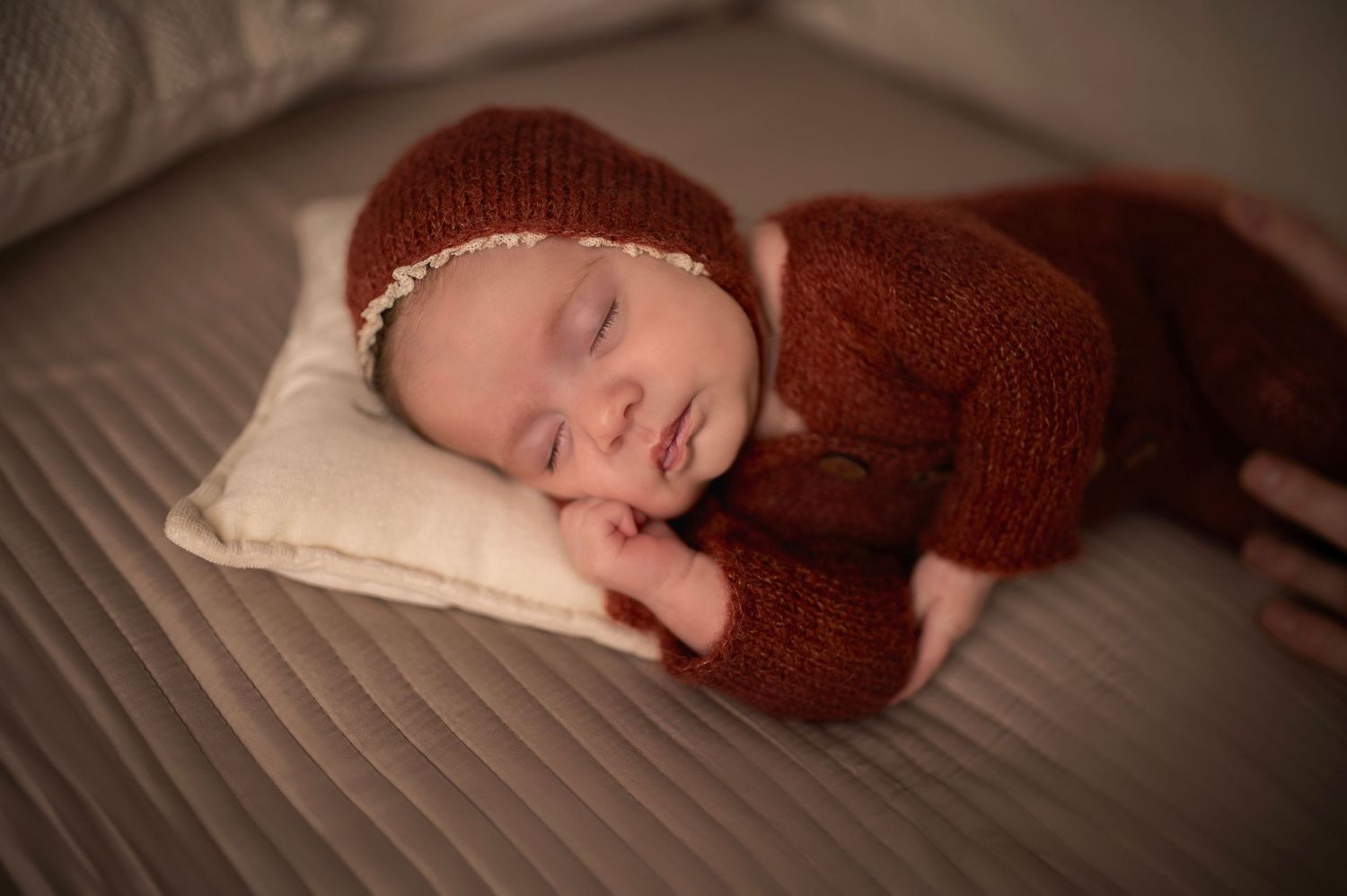 Sleeping baby in maroon knitted outfit and cap resting on white pillow.