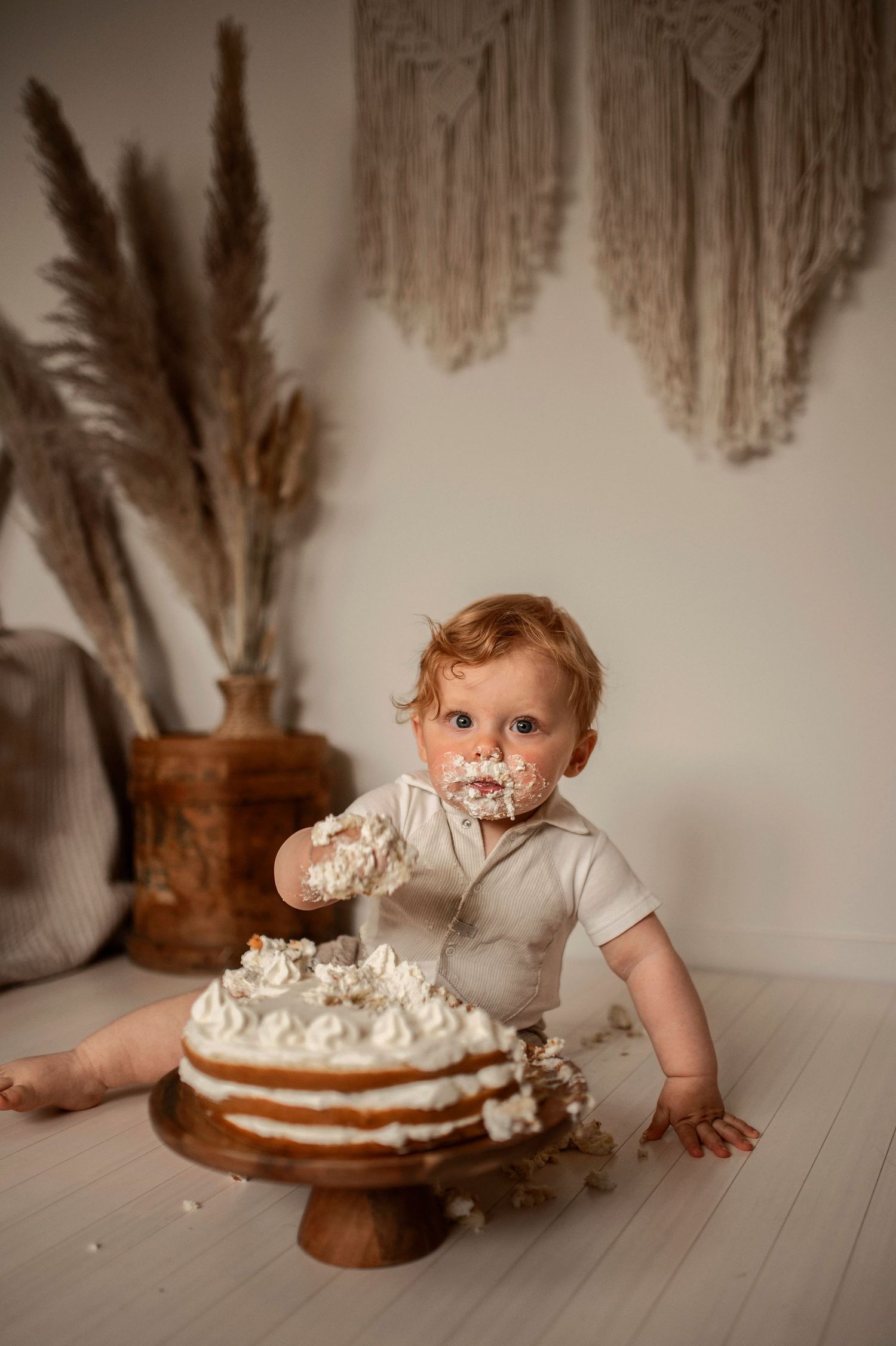 A baby enjoys a white frosted cake on a wooden stand by a macrame wall hanging and dried pampas grass decor.