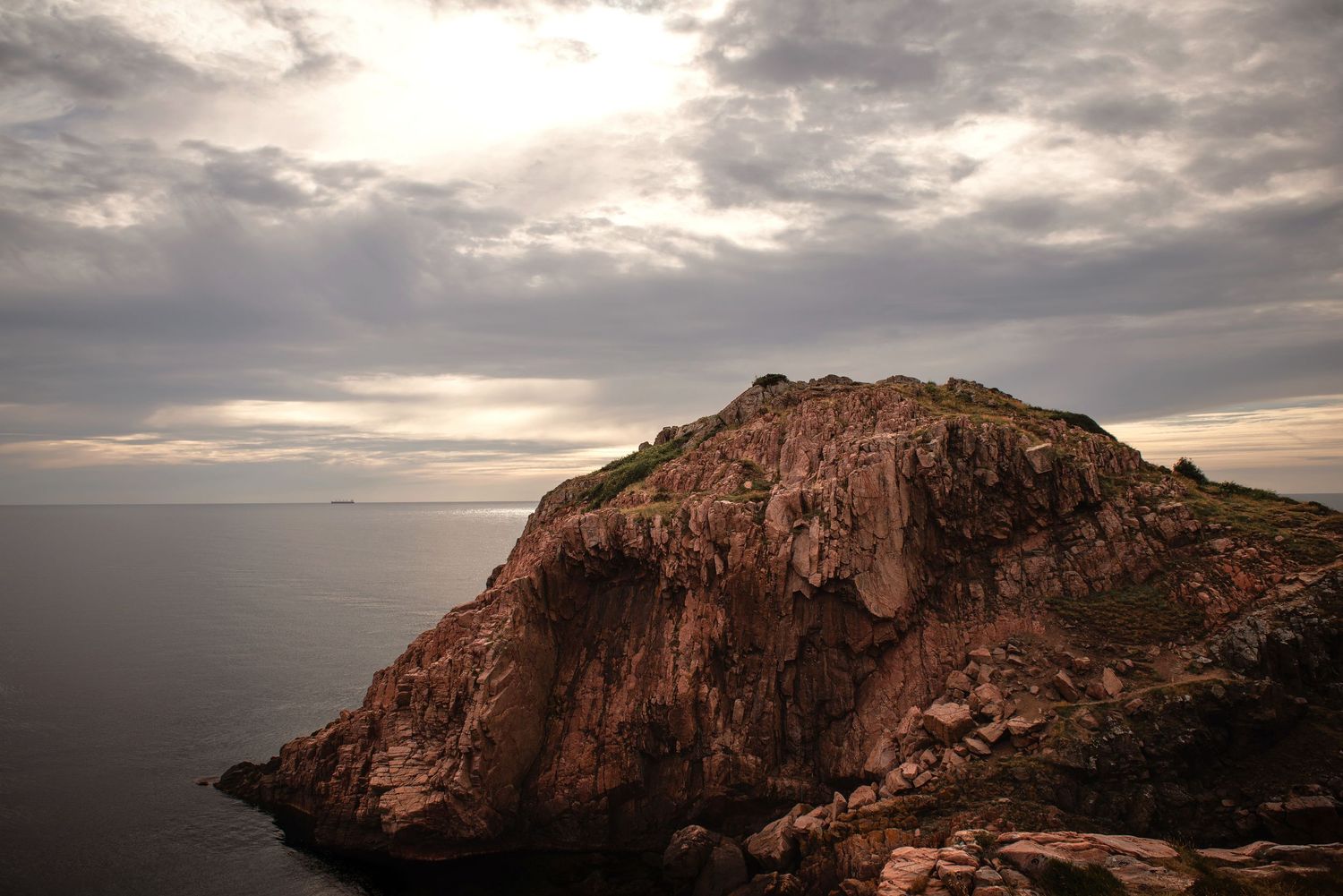 Rocky coastal cliff with green summit rises dramatically from calm sea under dramatic cloudy sky at dusk.