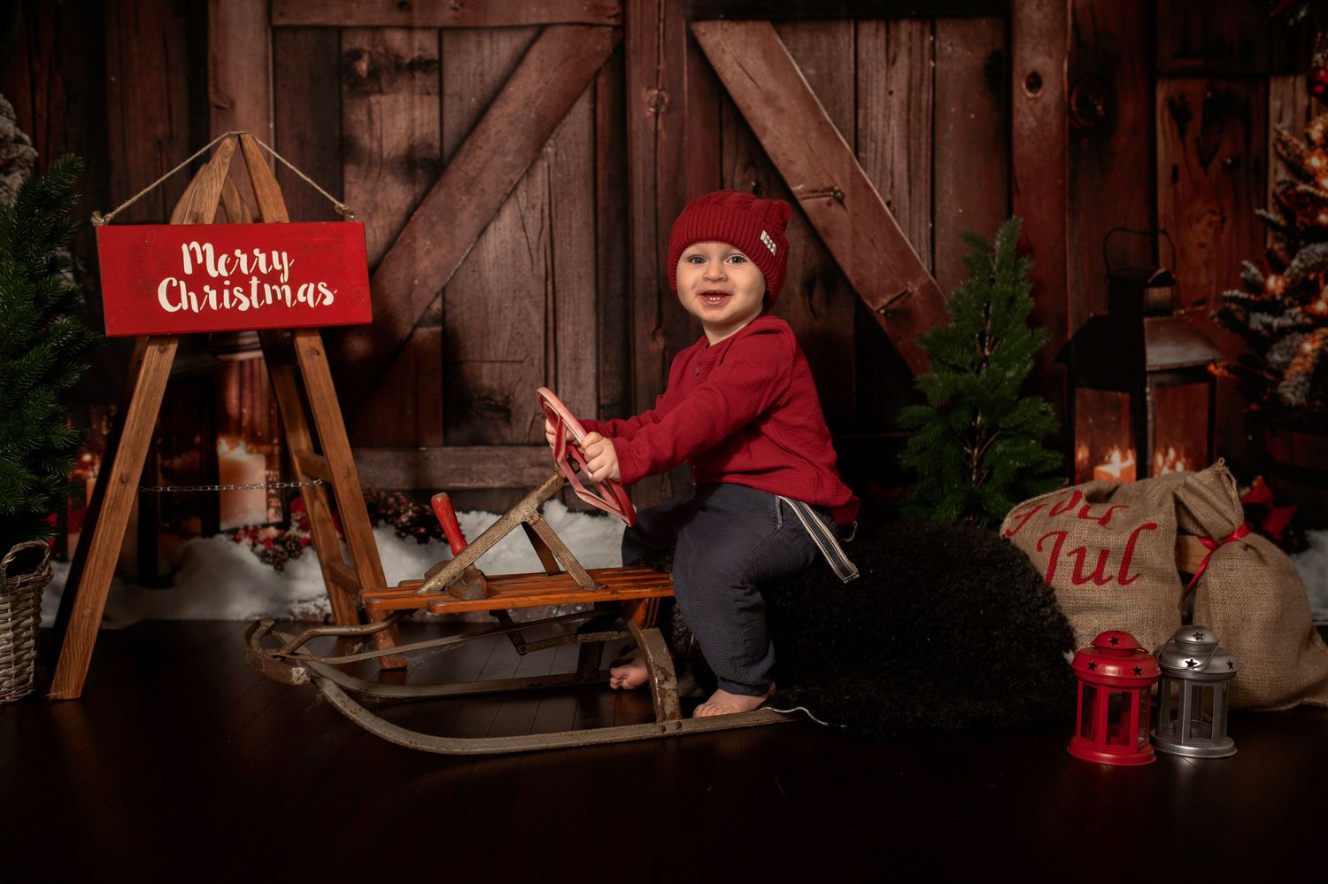 Child in red sweater and hat sits on vintage sled near Merry Christmas sign with rustic barn backdrop.