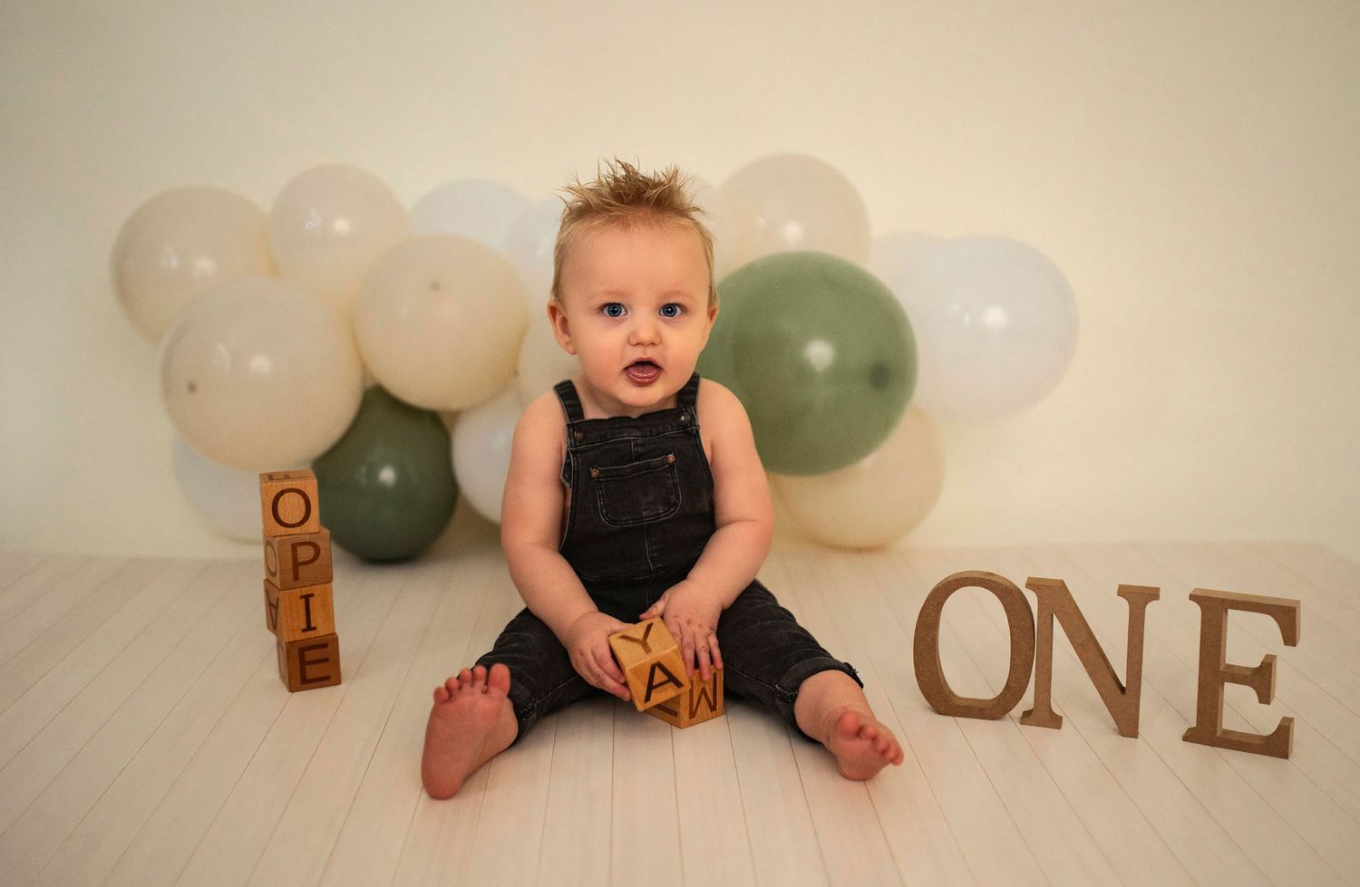 A first birthday photoshoot setup with white and sage green balloons and wooden 'ONE' lettering.