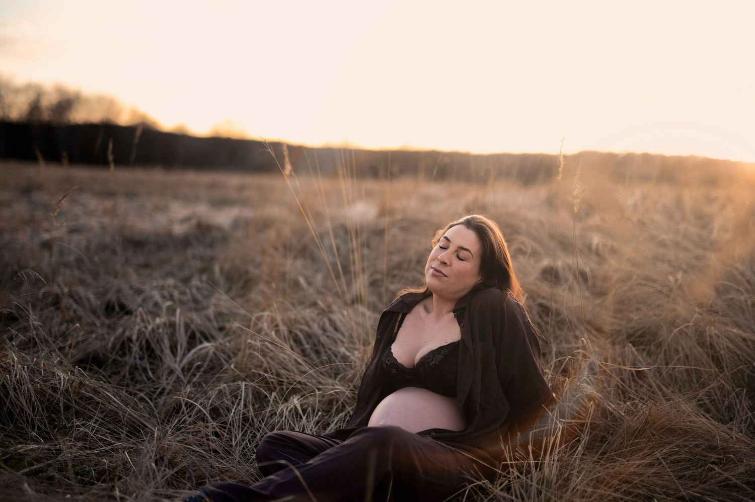 Pregnant woman sitting in golden field at sunset wearing dark cardigan.