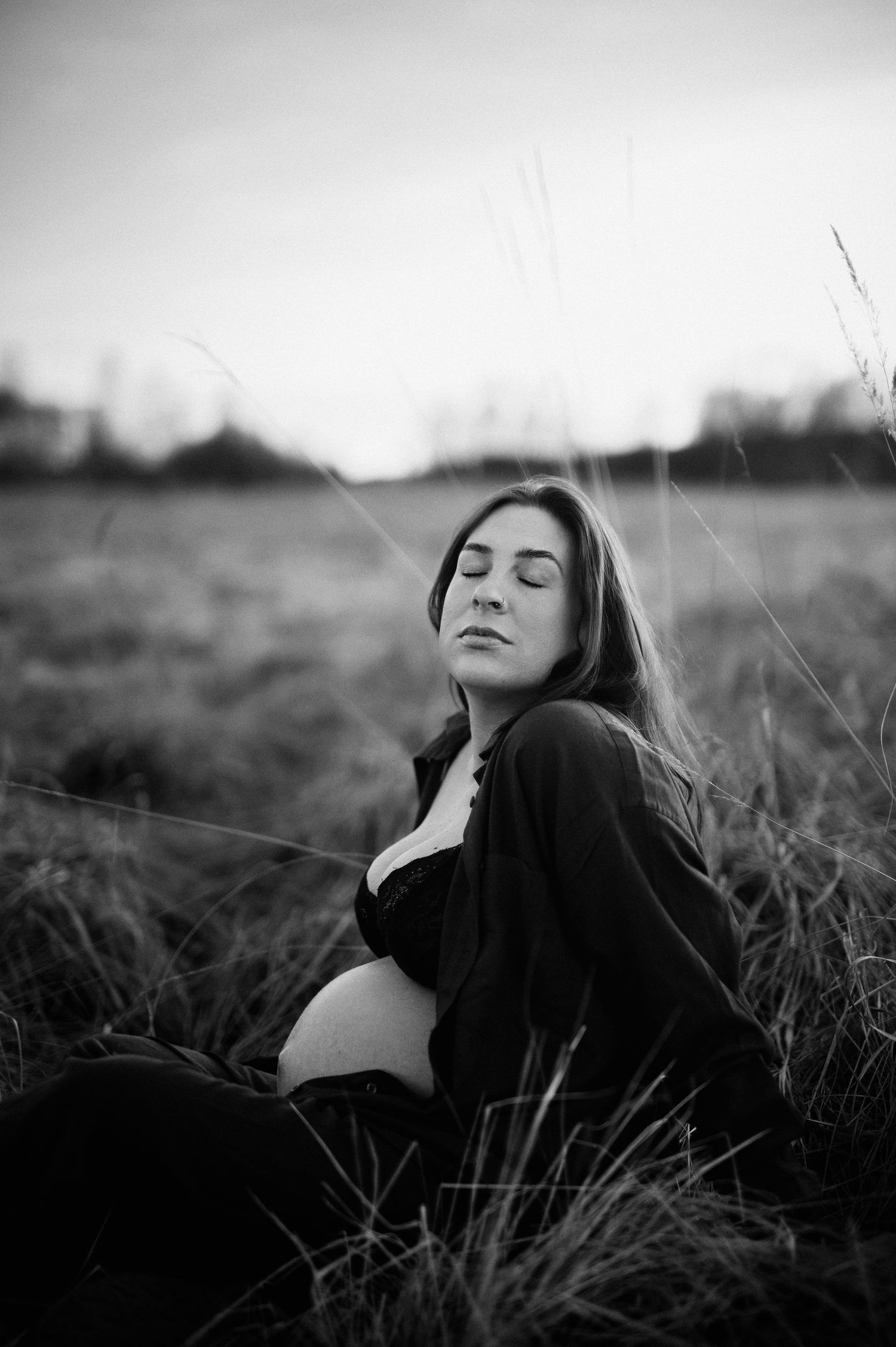 Svartvit gravidfotografering i Skåne med gravid kvinna i högt gräs i naturen i mjukt kvällsljus.