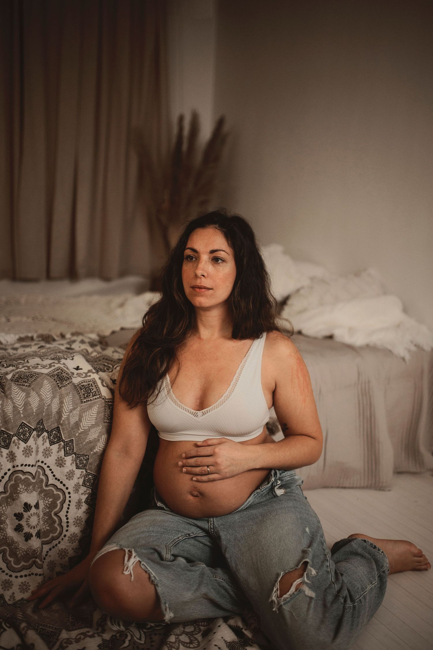 Pregnant woman in white crop top and ripped jeans sitting on floor beside bed, cradling baby bump.