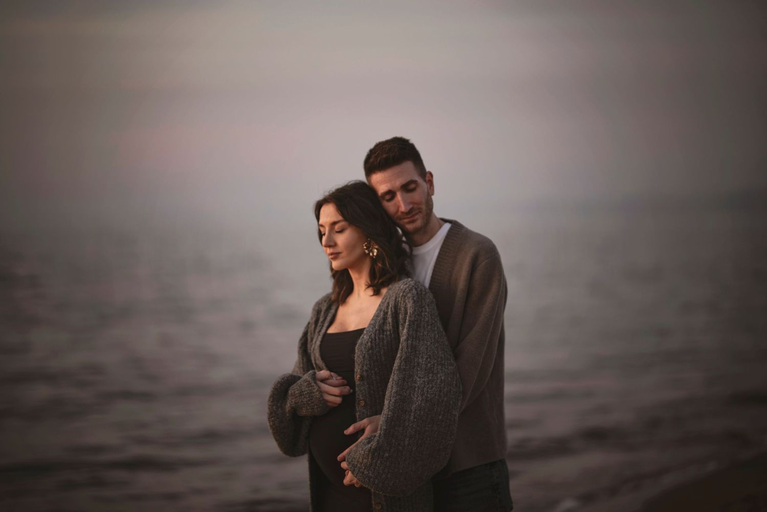 Romantiskt par omfamnas på en dimmig strand i solnedgången – en stämningsfull gravidfotografering i Skåne nära Hässleholm.
