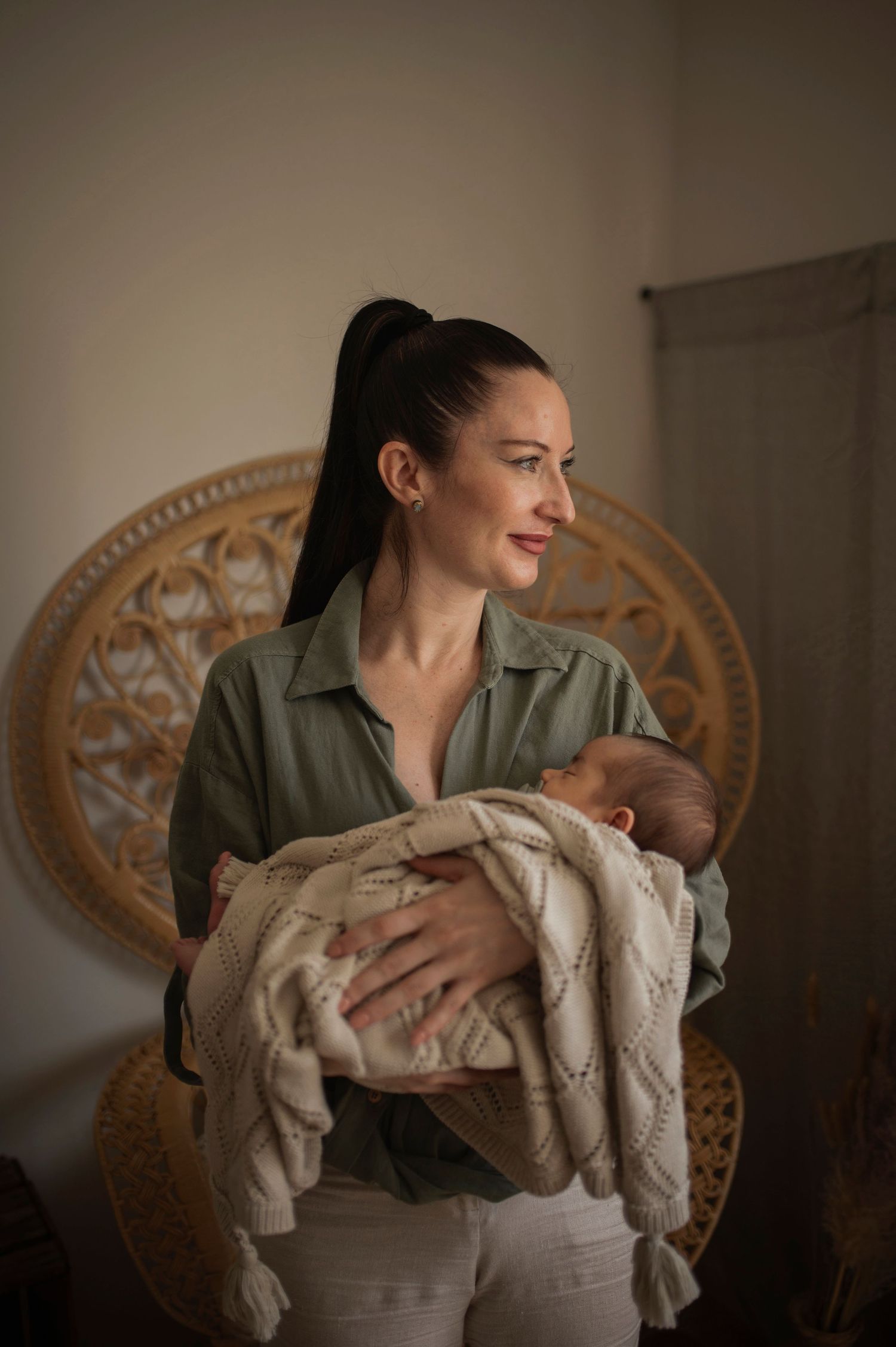 Parent in green shirt holding newborn in woven chair against neutral wall.