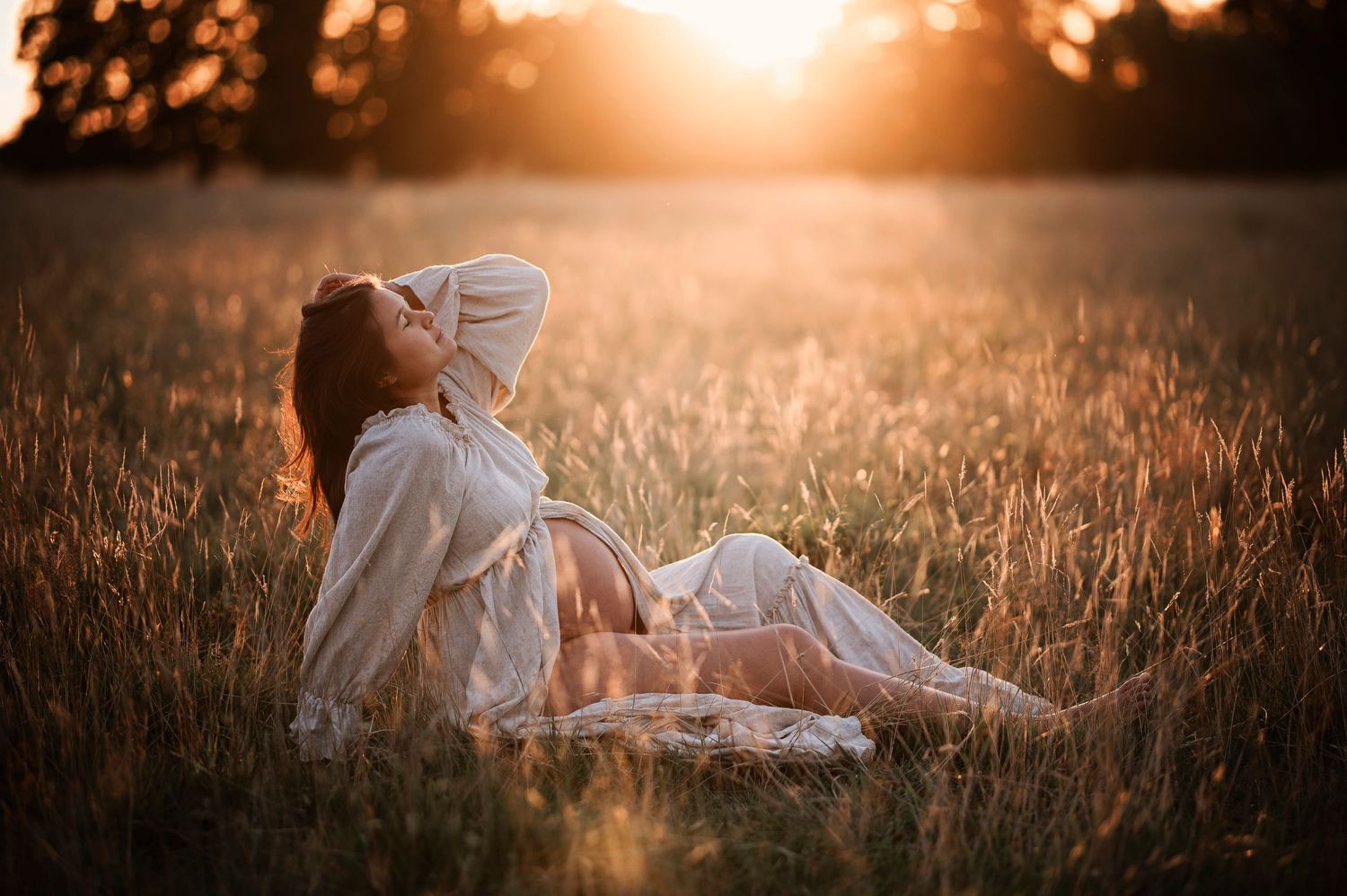 Expectant mother in white dress reclines in sunlit field during golden hour.