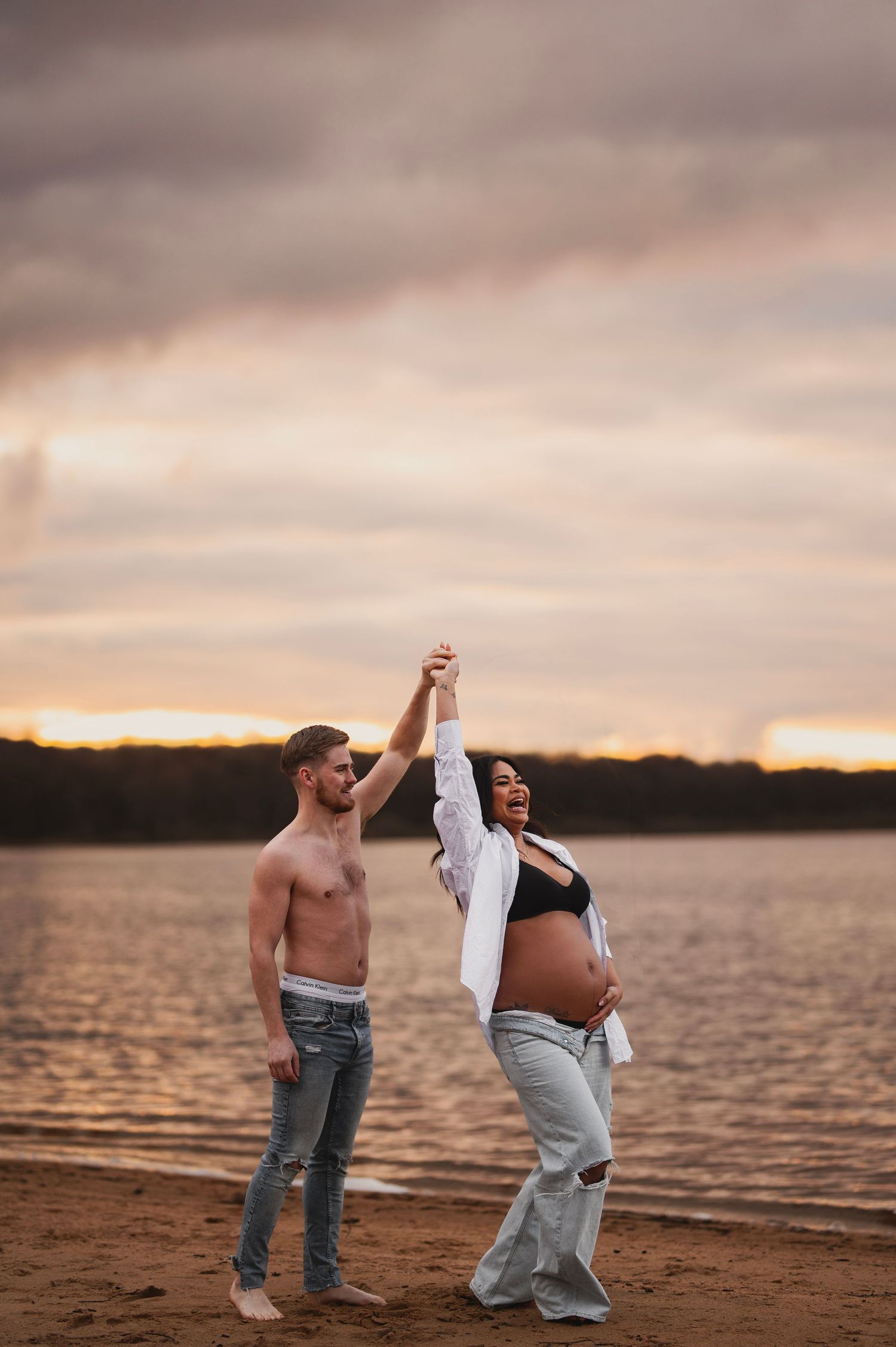 Couple dancing joyfully on beach during sunset maternity photoshoot.