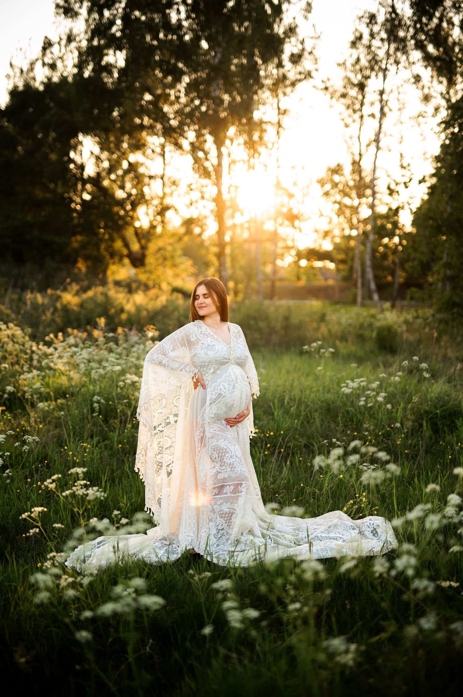 Gravidfotografering i Skåne med blivande mamma i vit spetsklänning i blomsteräng med vildmorot i solnedgång.
