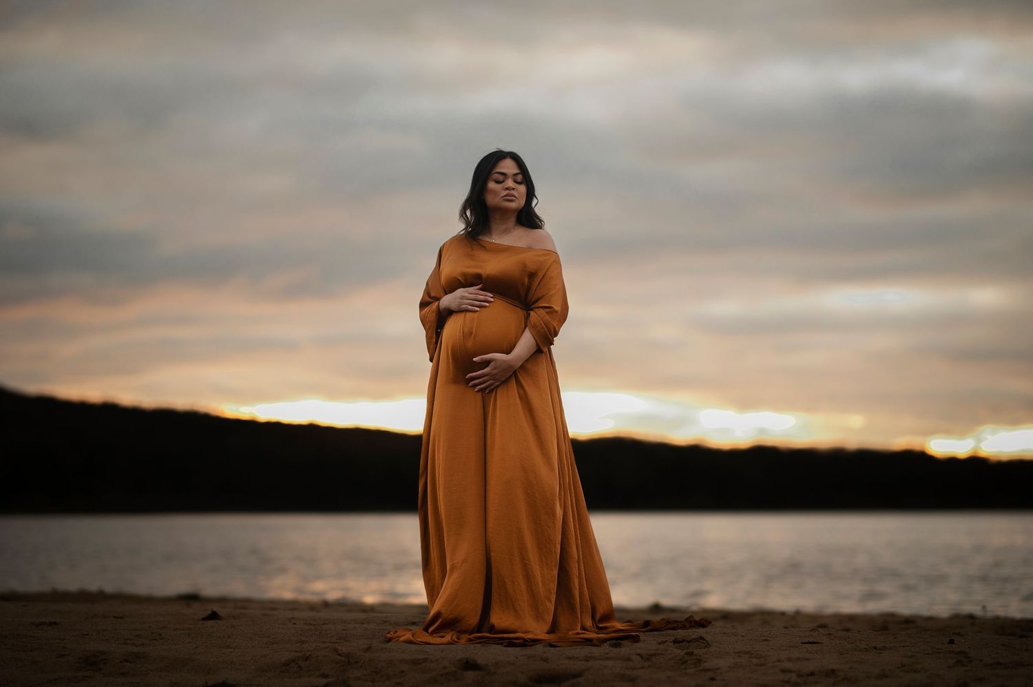 An expectant mother in a flowing rust-colored dress poses against a sunset lake backdrop.