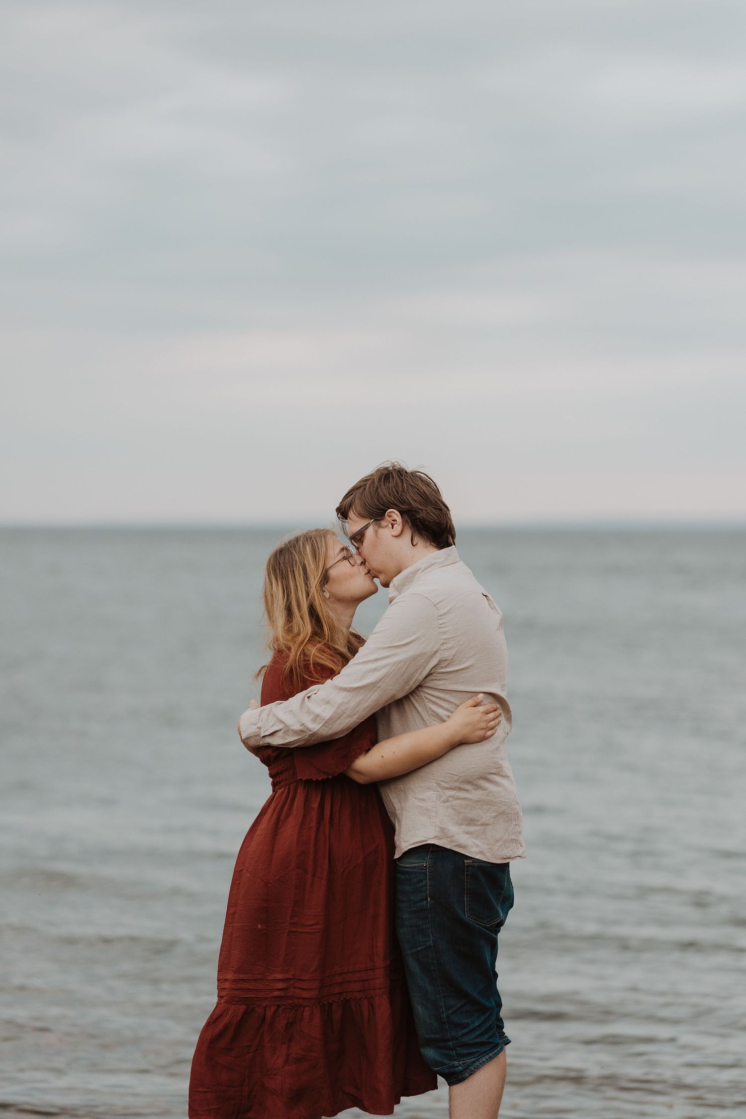 Couple sharing a romantic kiss on a beach, woman in red dress embracing man in casual attire with ocean backdrop at Hovs Hallar