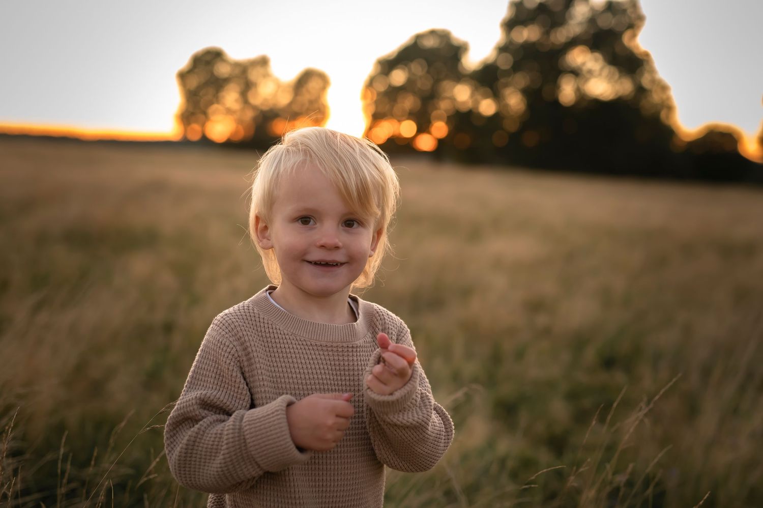Barnfotografering i Skåne med leende pojke i beige tröja i gyllene fält i solnedgång.