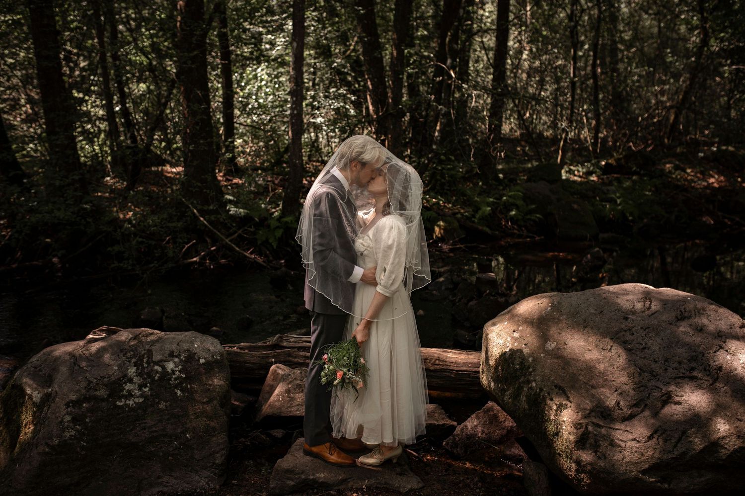 Ett par delar en romantisk kyss under en spetsslöja i en lummig skogsmiljö på sin bröllopsdag i Skåne