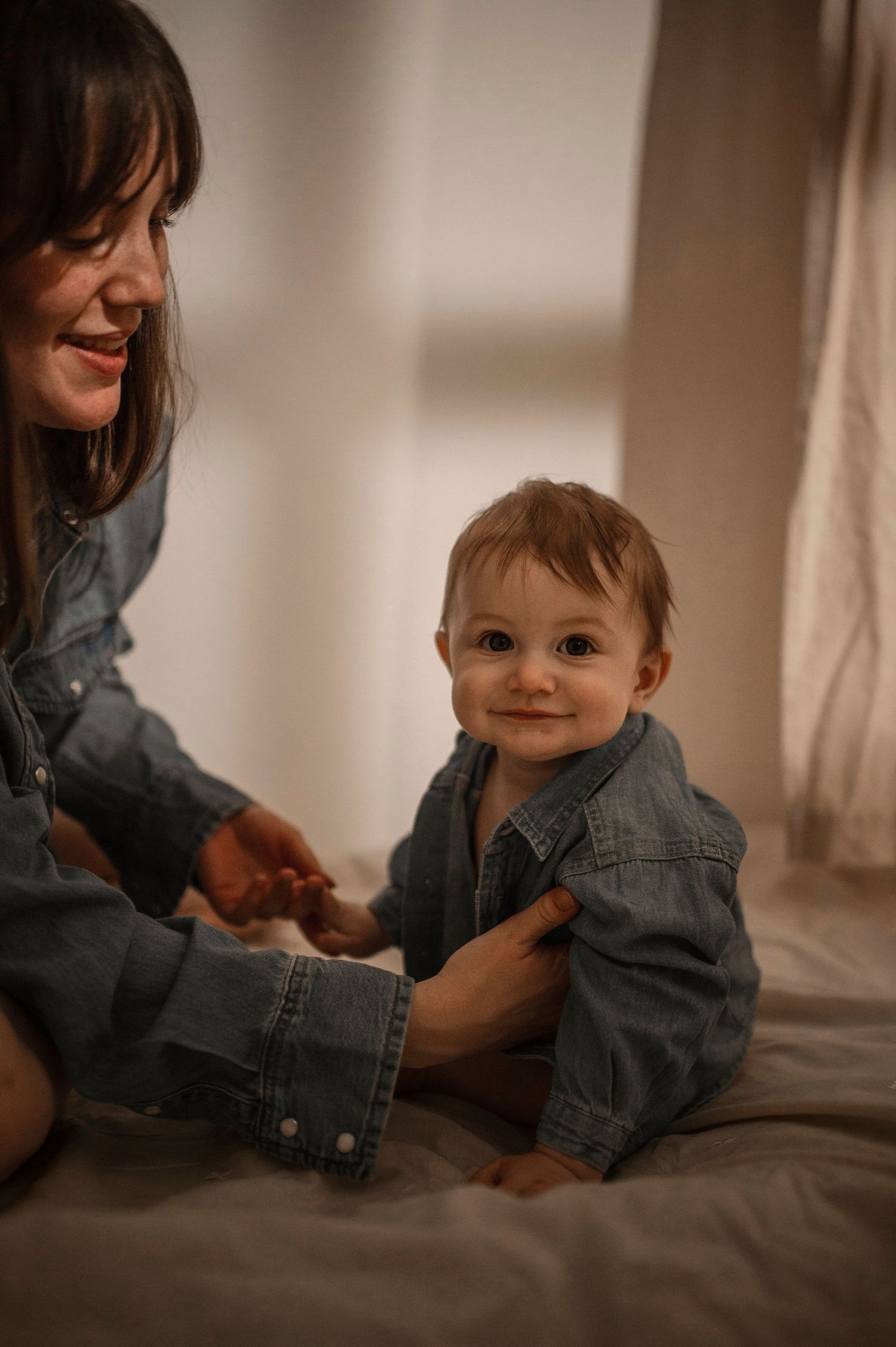 A smiling baby in a denim shirt sits on a bed while a woman in matching denim lovingly holds their hands.