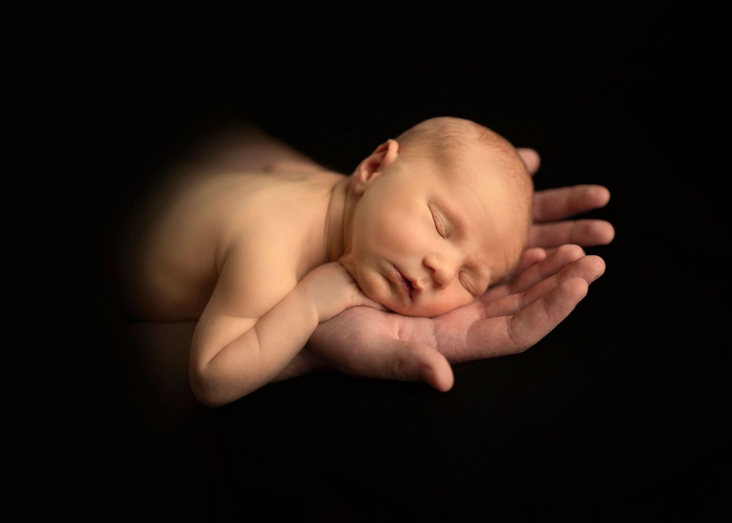 A peaceful newborn baby sleeps on dark background in artistic portrait pose.
