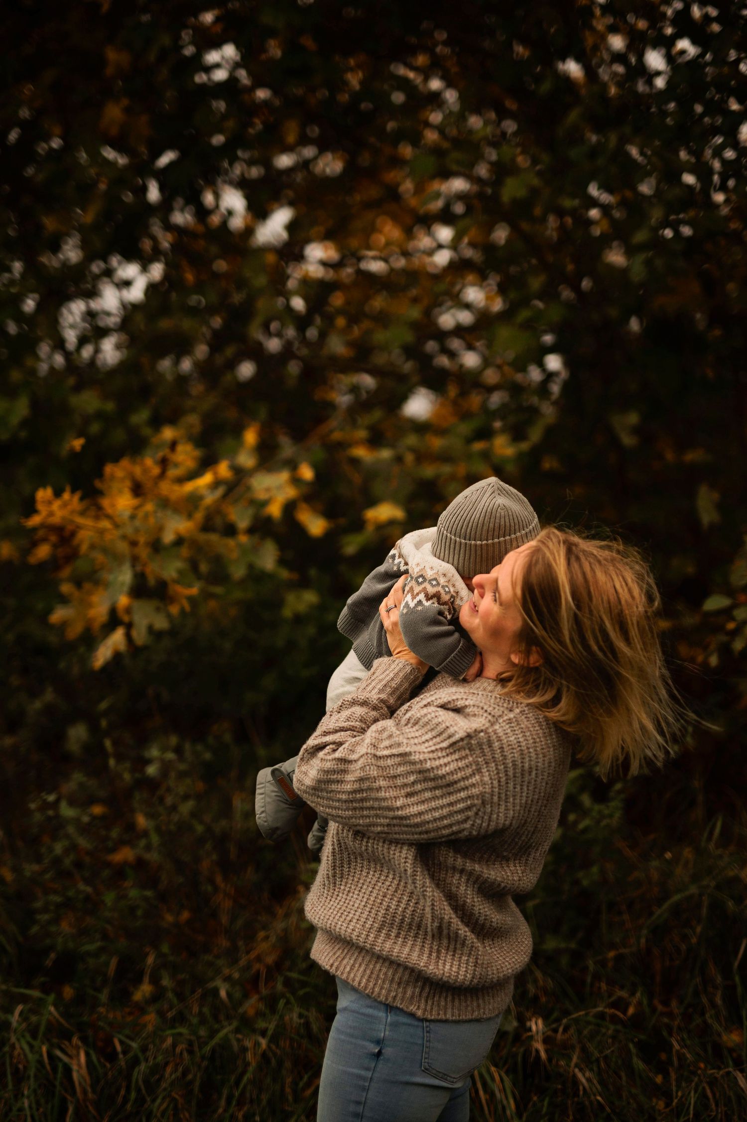 A candid outdoor moment of parent and baby against autumn trees.