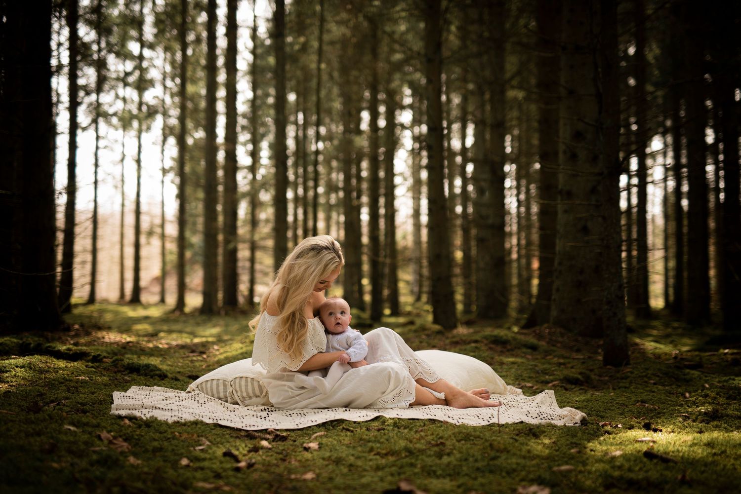Mother and baby resting on blanket in sunlit forest clearing.