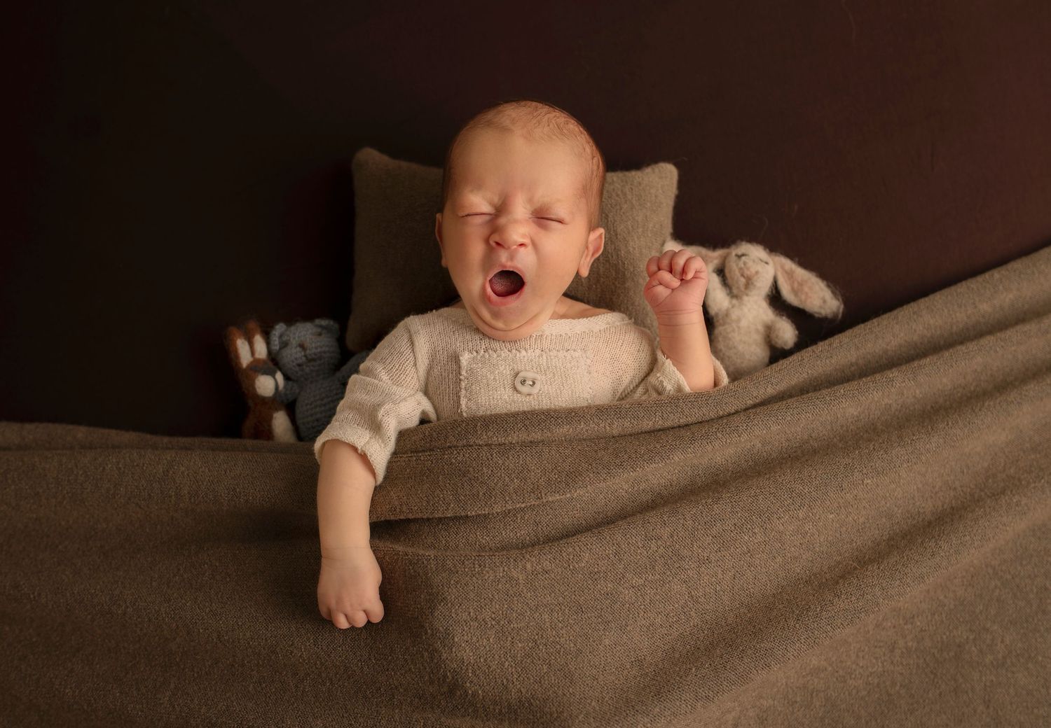 Baby in white onesie yawning while lying on brown blanket.
