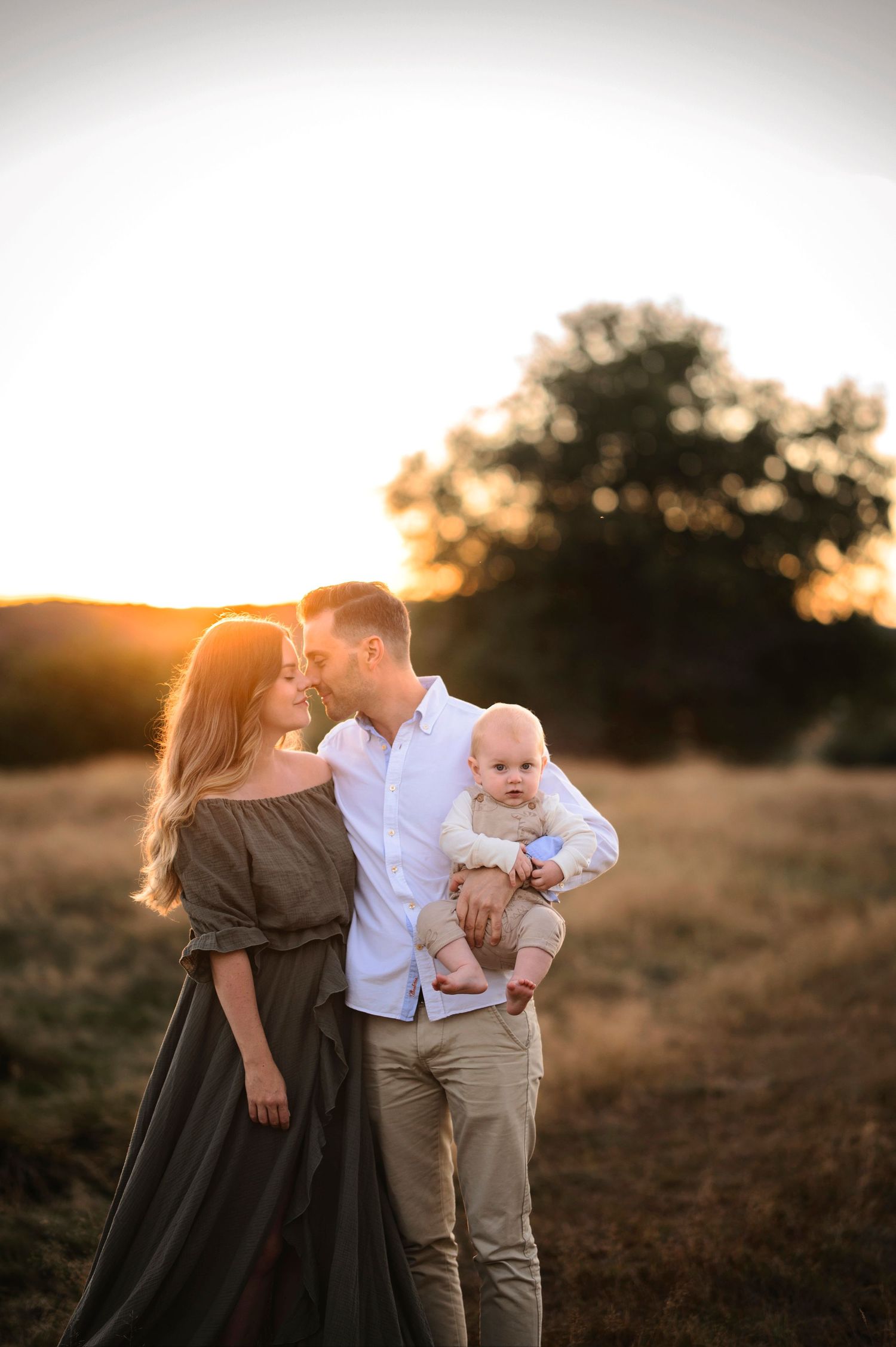 Familjefotografering i Skåne med föräldrar som håller sitt barn i gyllene solnedgång på fält.