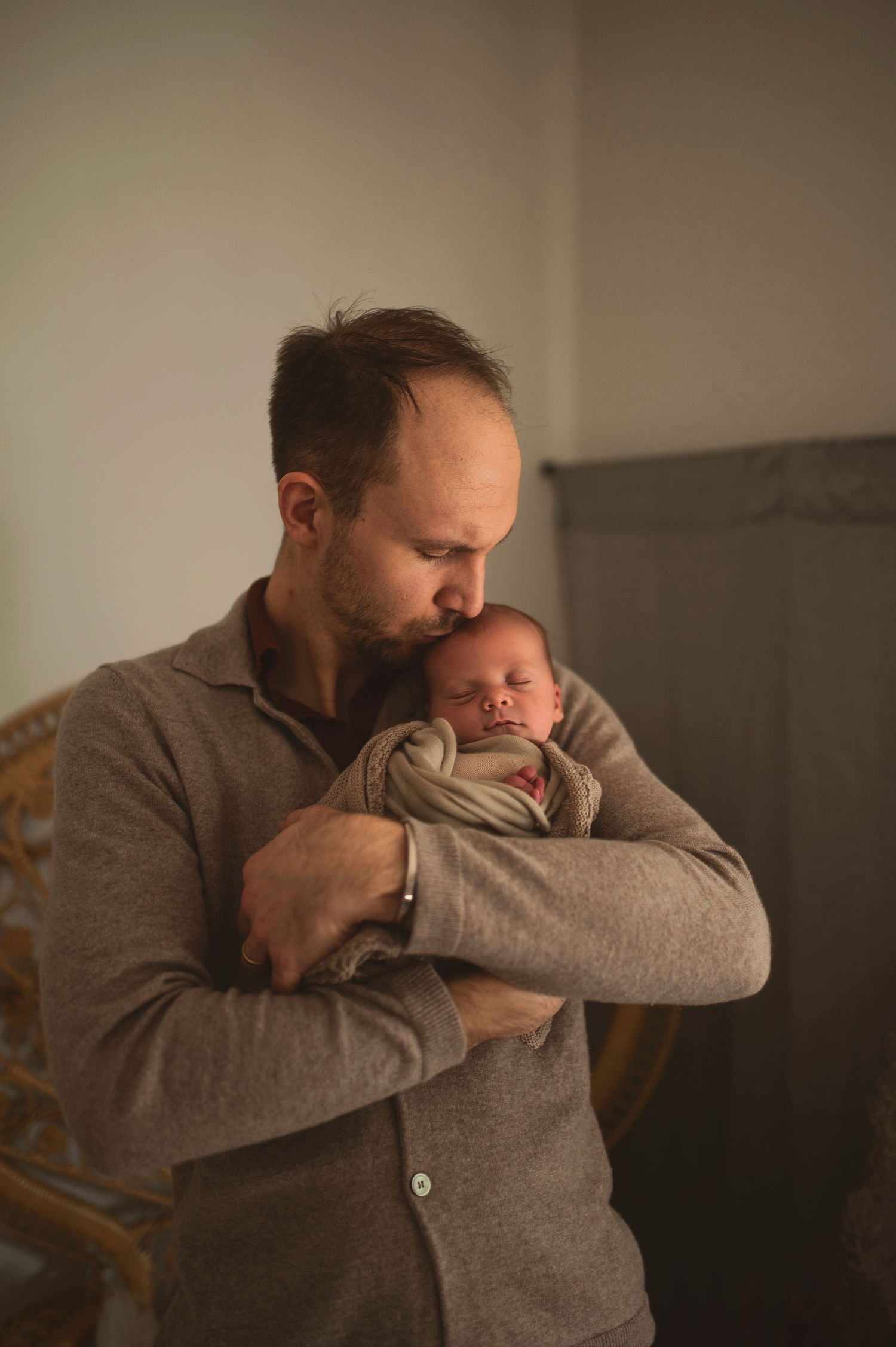 Parent in gray sweater holding sleeping newborn against warm neutral background.