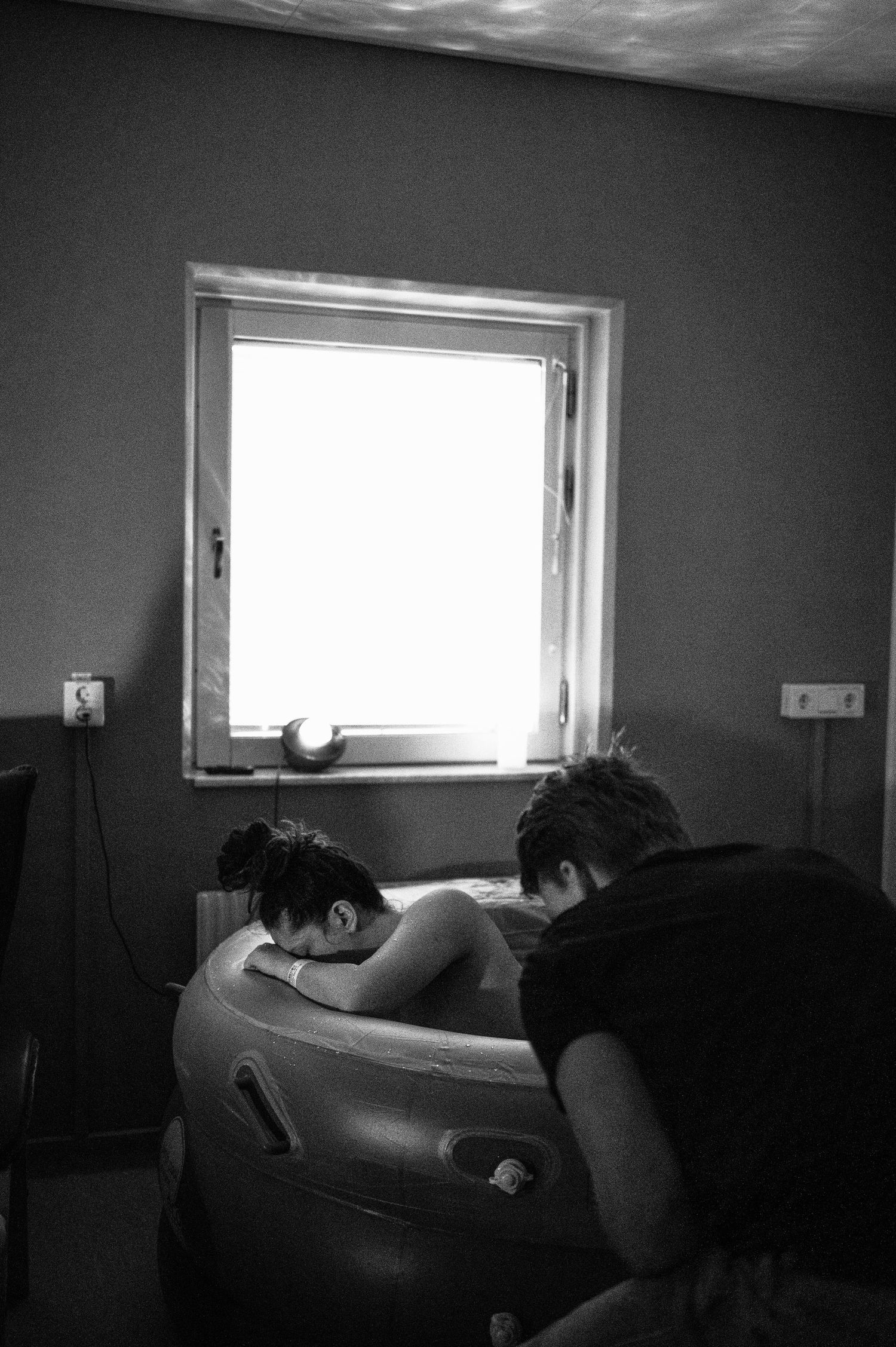 Black and white photo showing a person laboring in a birthing pool next to a bright window.