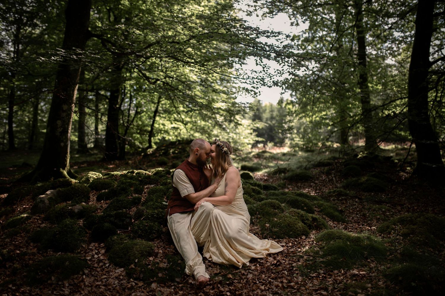 Bröllopsfotografering i Skåne med par i romantisk skogsmiljö sittande på mossklädda stenar.