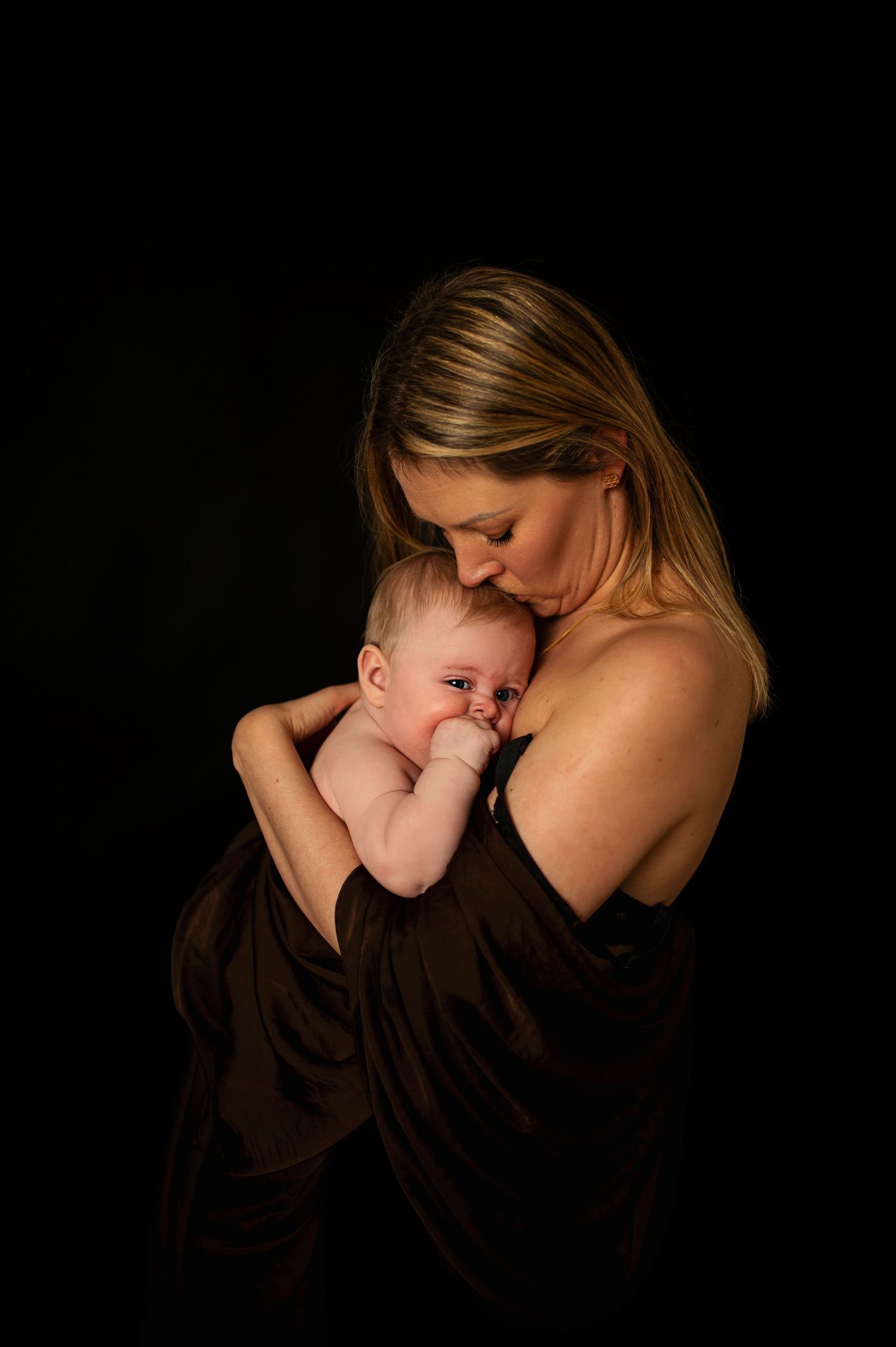 Intimate mother and baby portrait in dark dramatic lighting against black background.