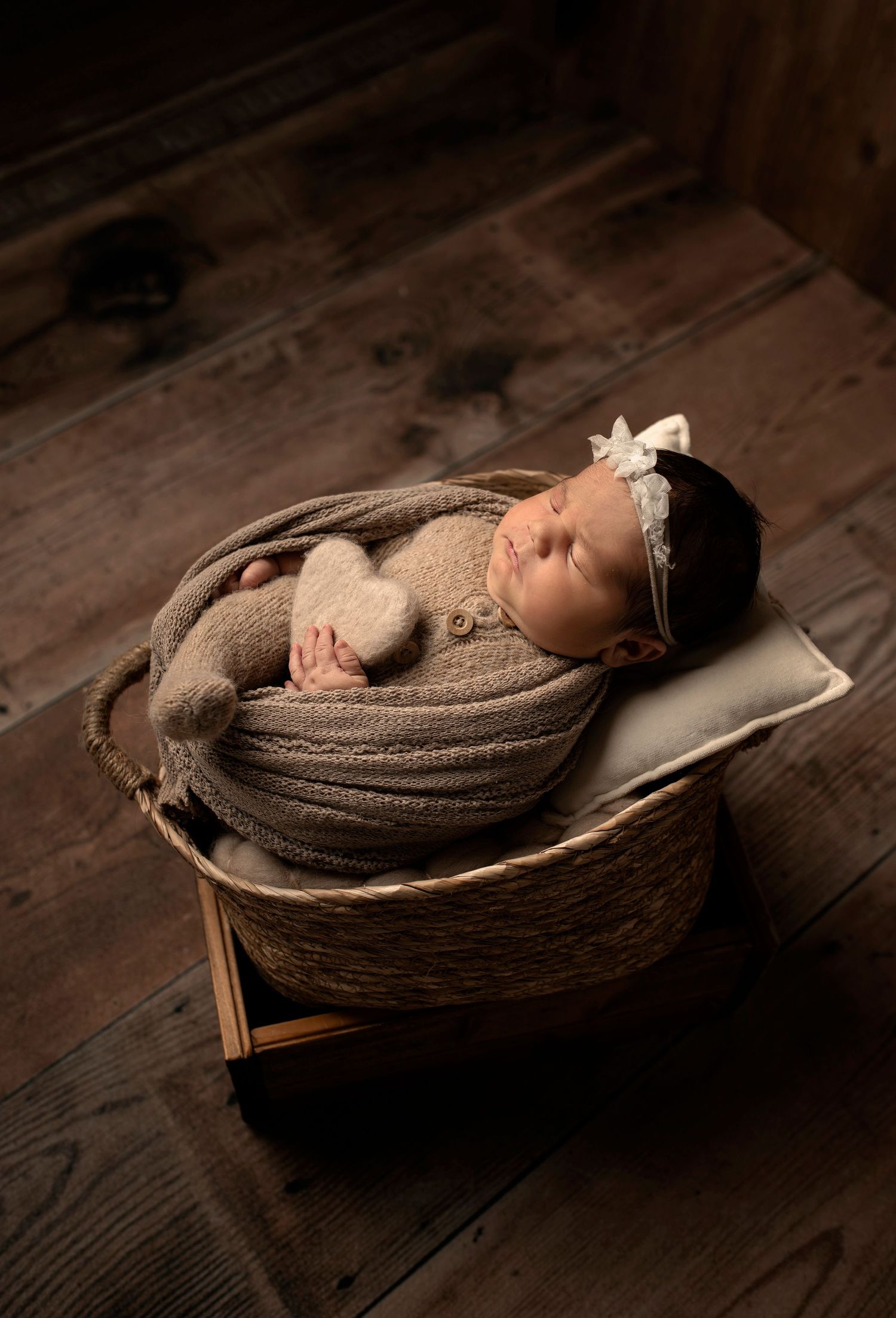 Sleeping newborn baby wrapped in brown knit blanket with bow headband in wicker basket prop.