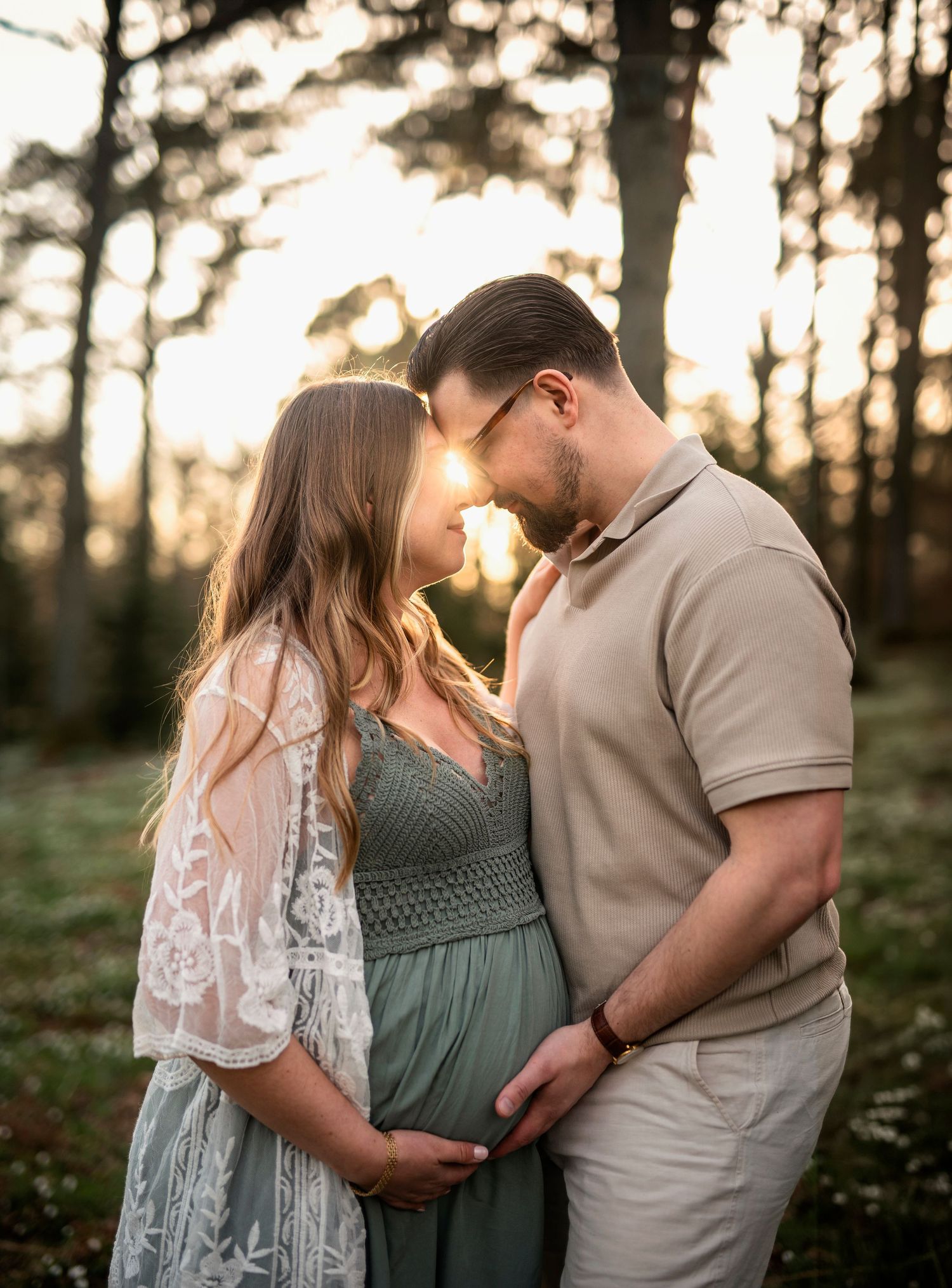 Expectant couple touching foreheads tenderly in sunlit forest clearing.