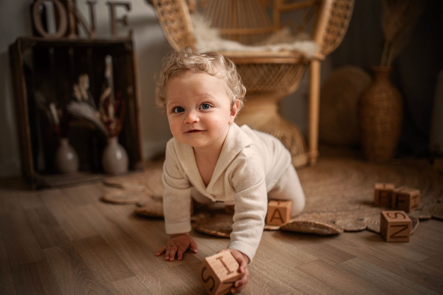 Baby in cream sweater crawls on wooden floor near vintage decor and wicker furniture.