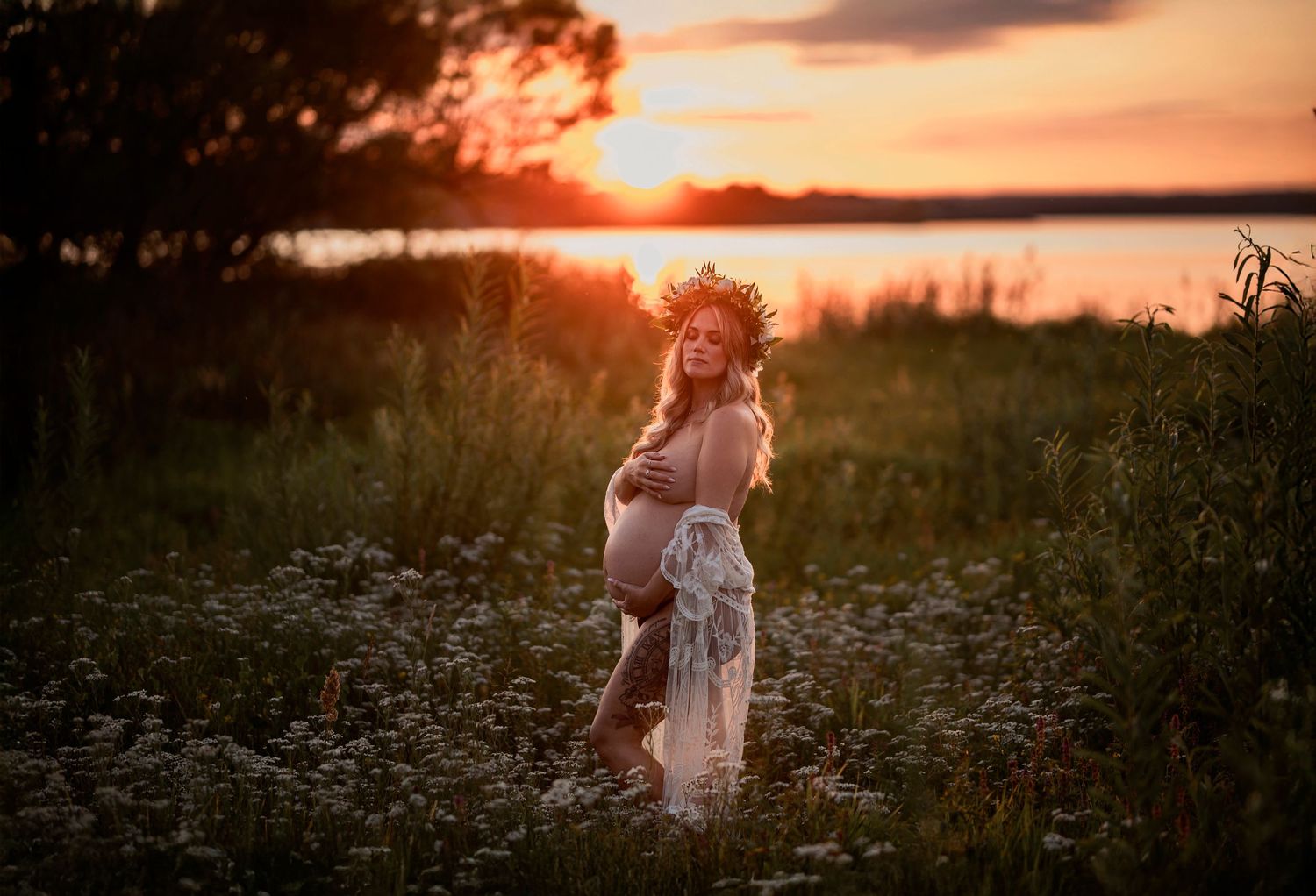 Pregnant woman in floral crown stands in a meadow during a vibrant golden sunset.