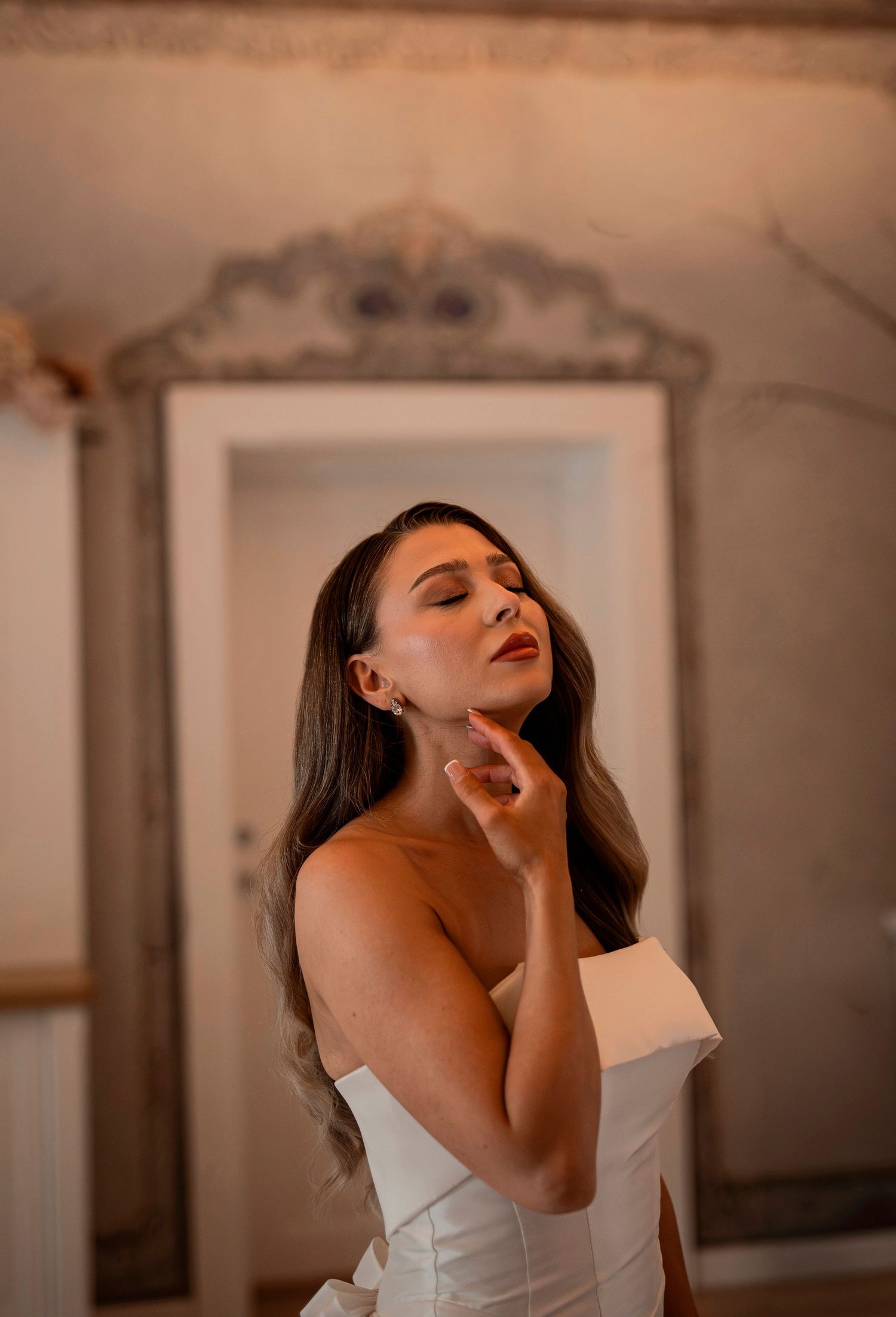 Elegant bride in white off-shoulder gown poses gracefully before ornate vintage mirror with warm ambient lighting.