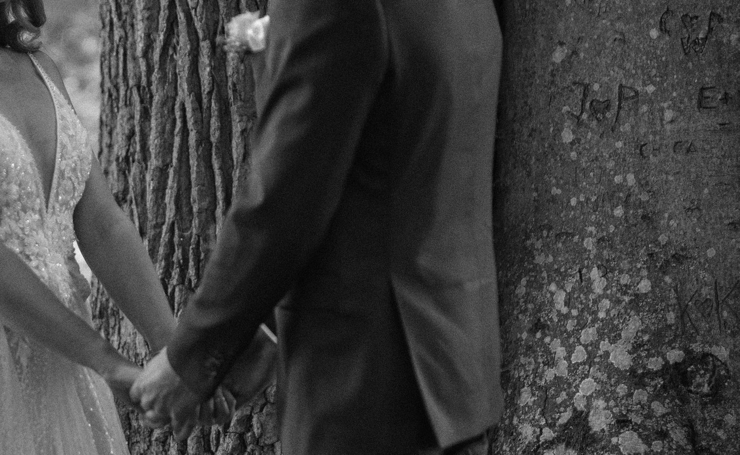 Black and white wedding photography in Skåne with bride and groom holding hands near a tree with carved initials during their wedding day.