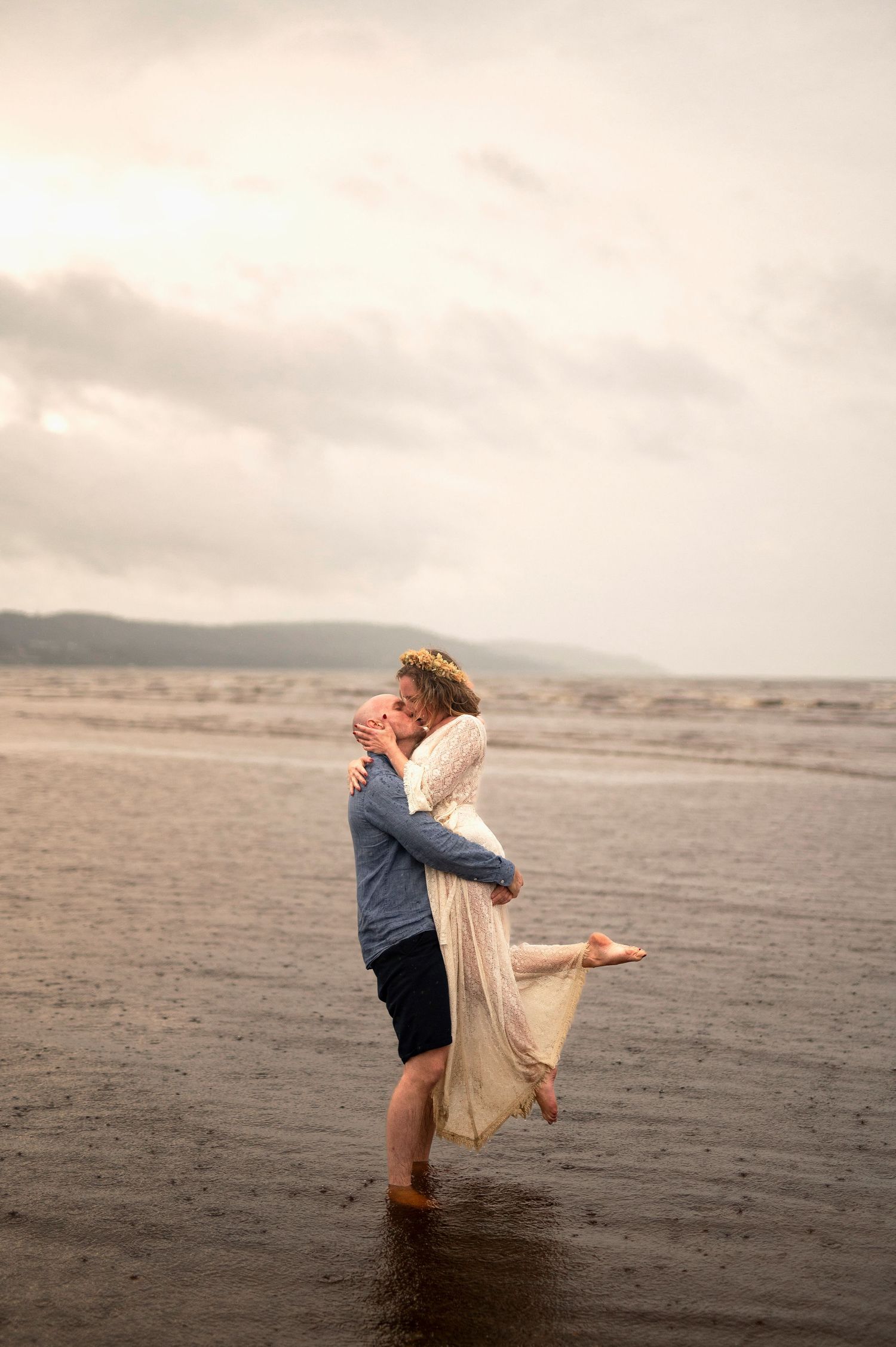 Couple embracing on a misty beach, man lifting woman in white dress with ocean waves in the background.