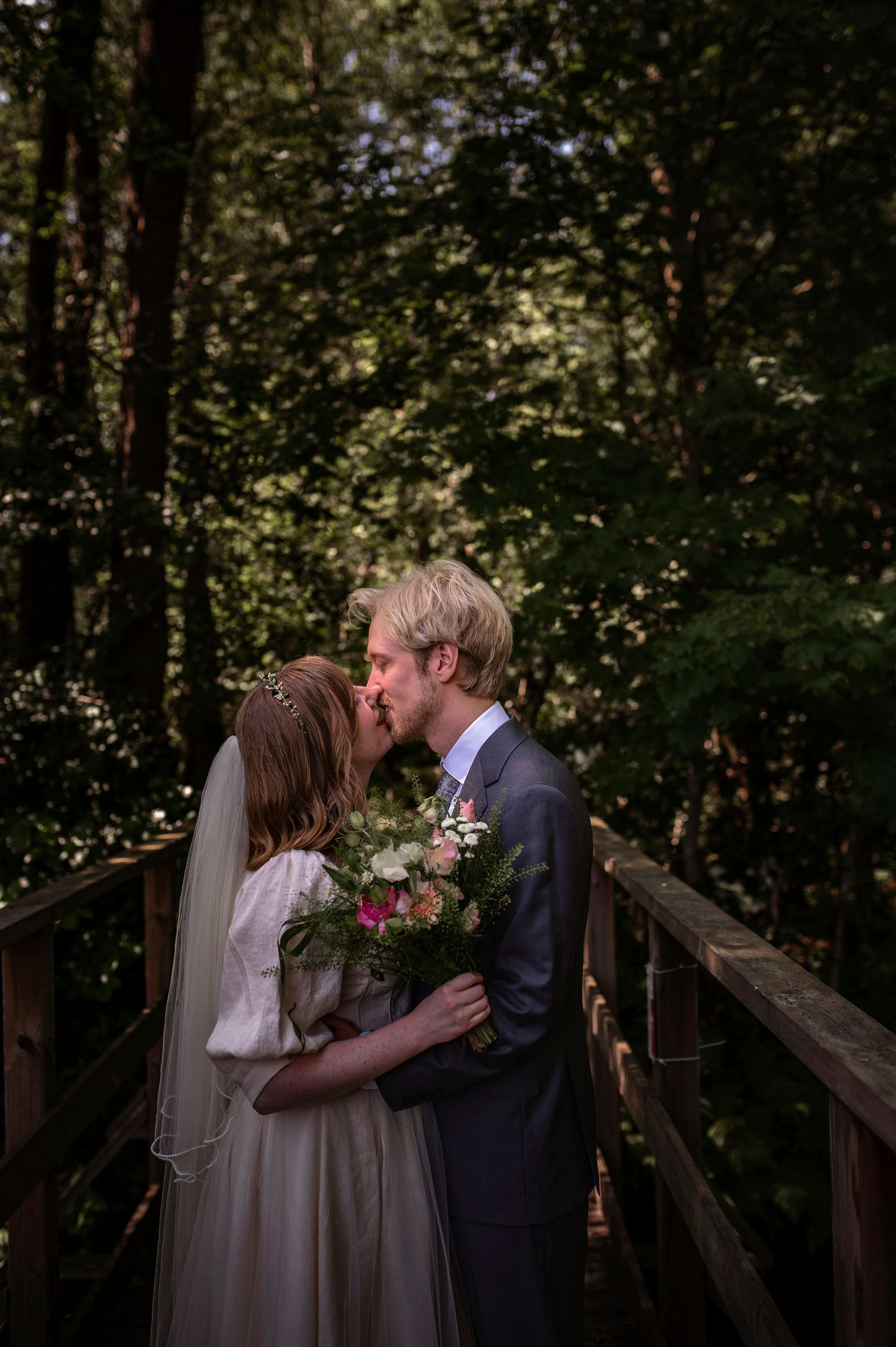 Bride and groom share a kiss on a wooden bridge surrounded by lush green forest trees on their wedding day.