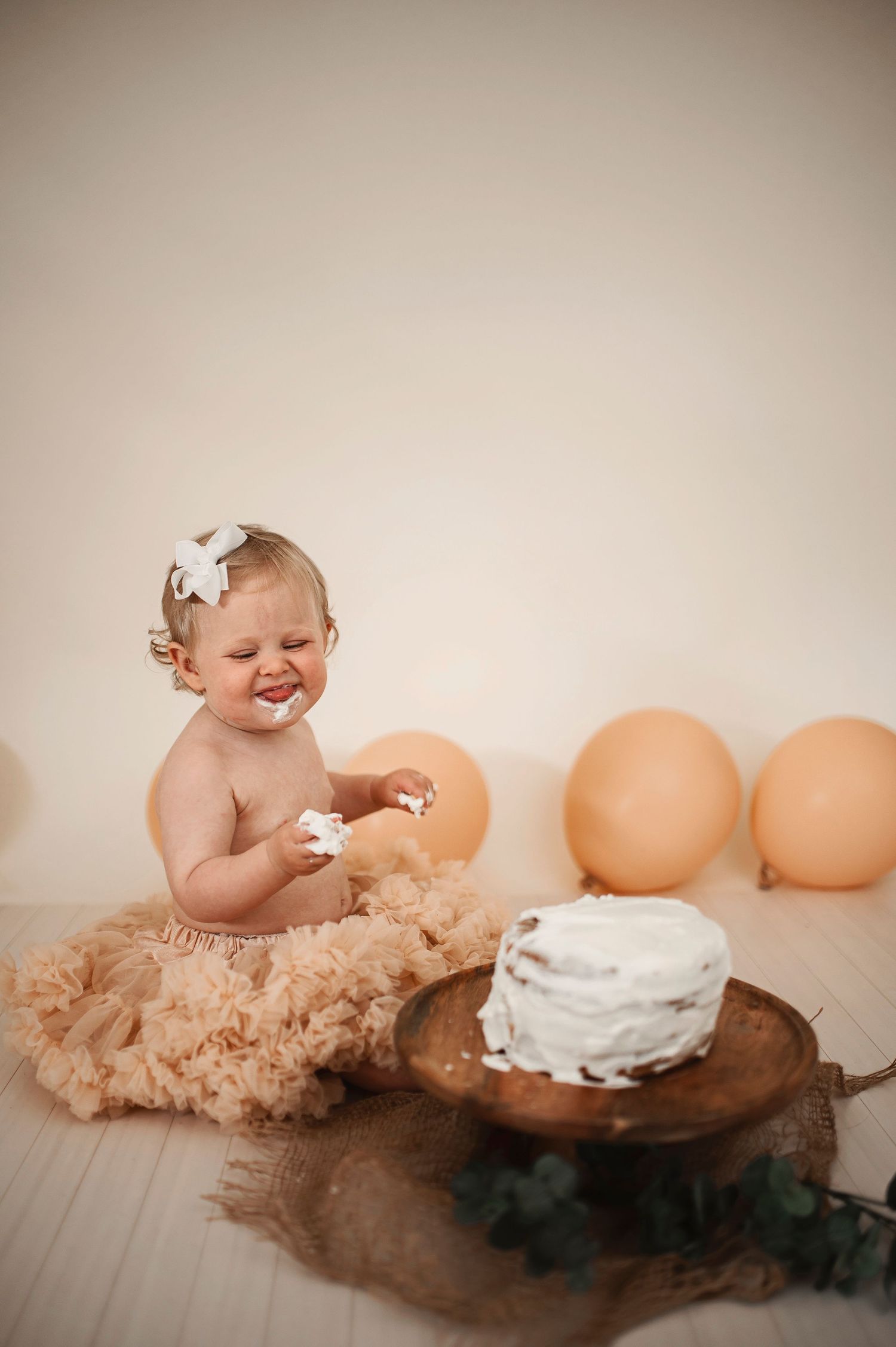 Happy baby with bow smiles while sitting next to birthday cake and peach balloons.