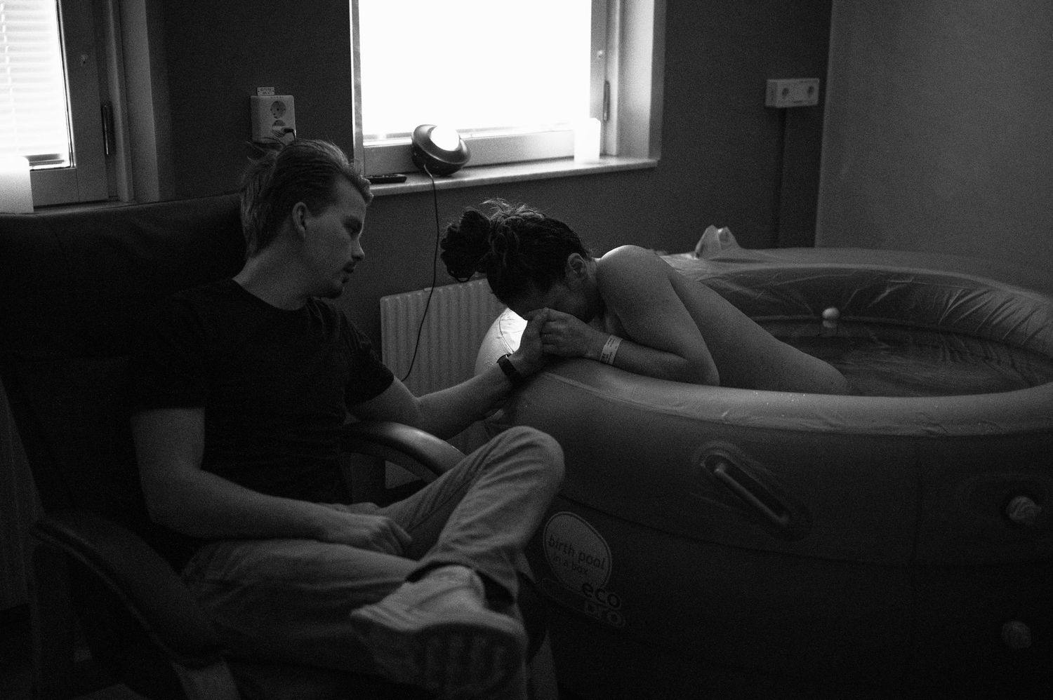 A black and white photograph of people sitting near an inflatable birthing pool in a dimly lit room.