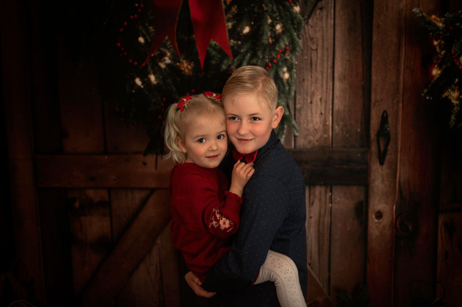 Two siblings pose for a Christmas portrait in front of a rustic wooden door with holiday wreath decor.