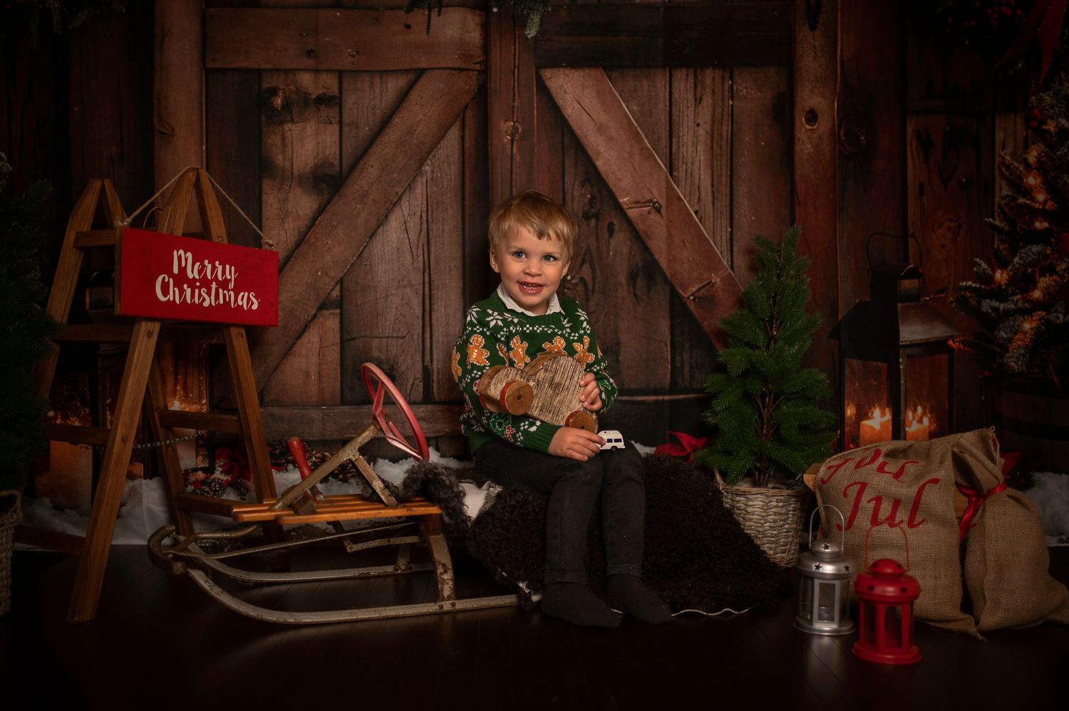A young boy sits on a vintage sled surrounded by Christmas decorations and a Merry Christmas sign in a rustic barn setting.