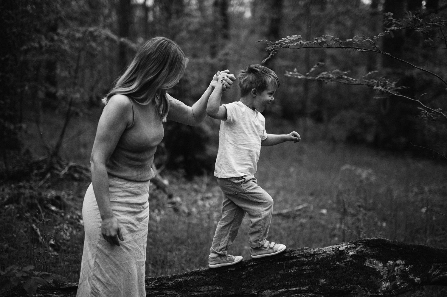 Familjefotografering i Skåne med mamma och son i lekfull balans på stock i skog i svartvitt ljus.
