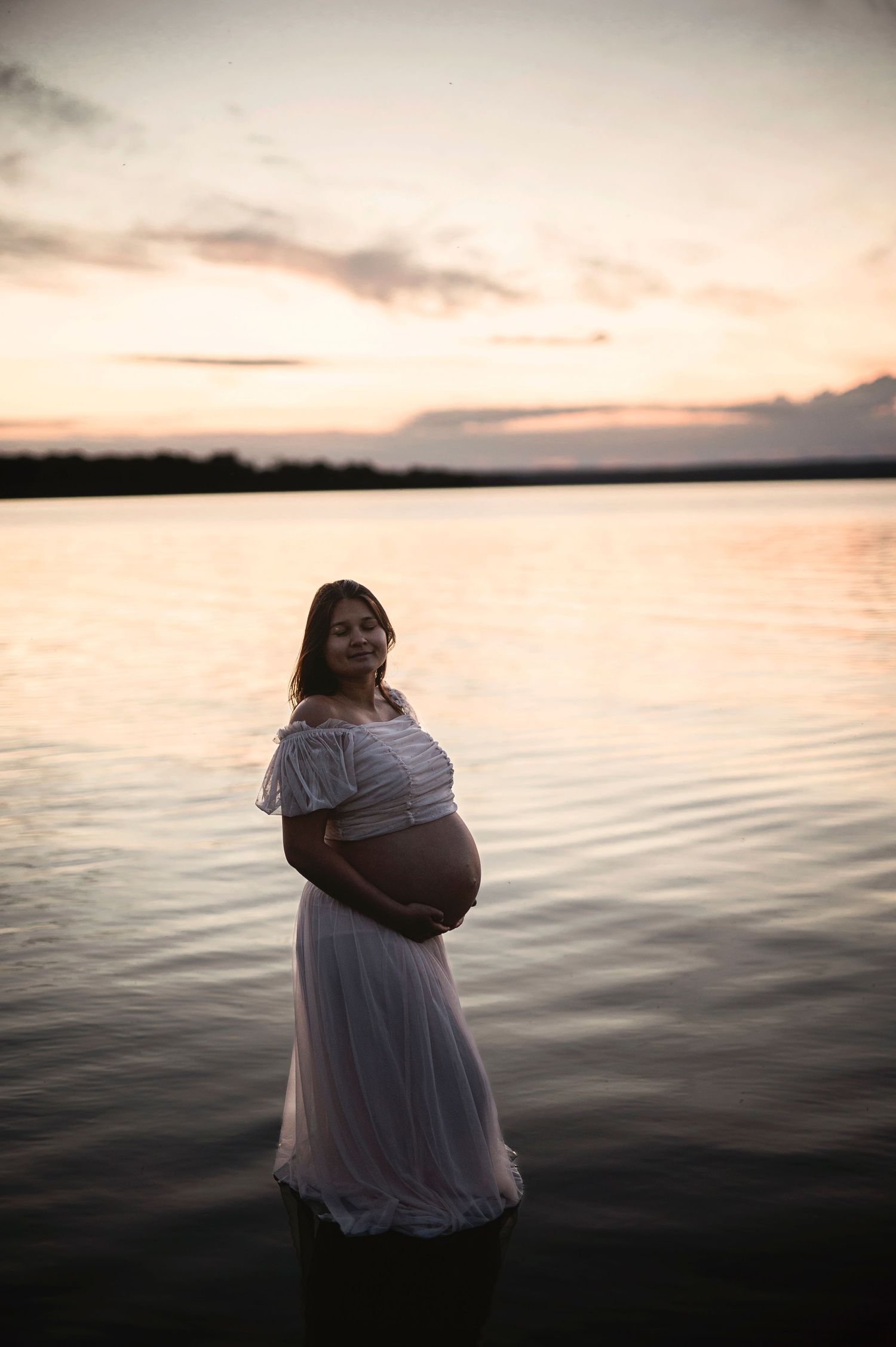 Artistic maternity portrait of woman in white dress standing in calm water at sunset.