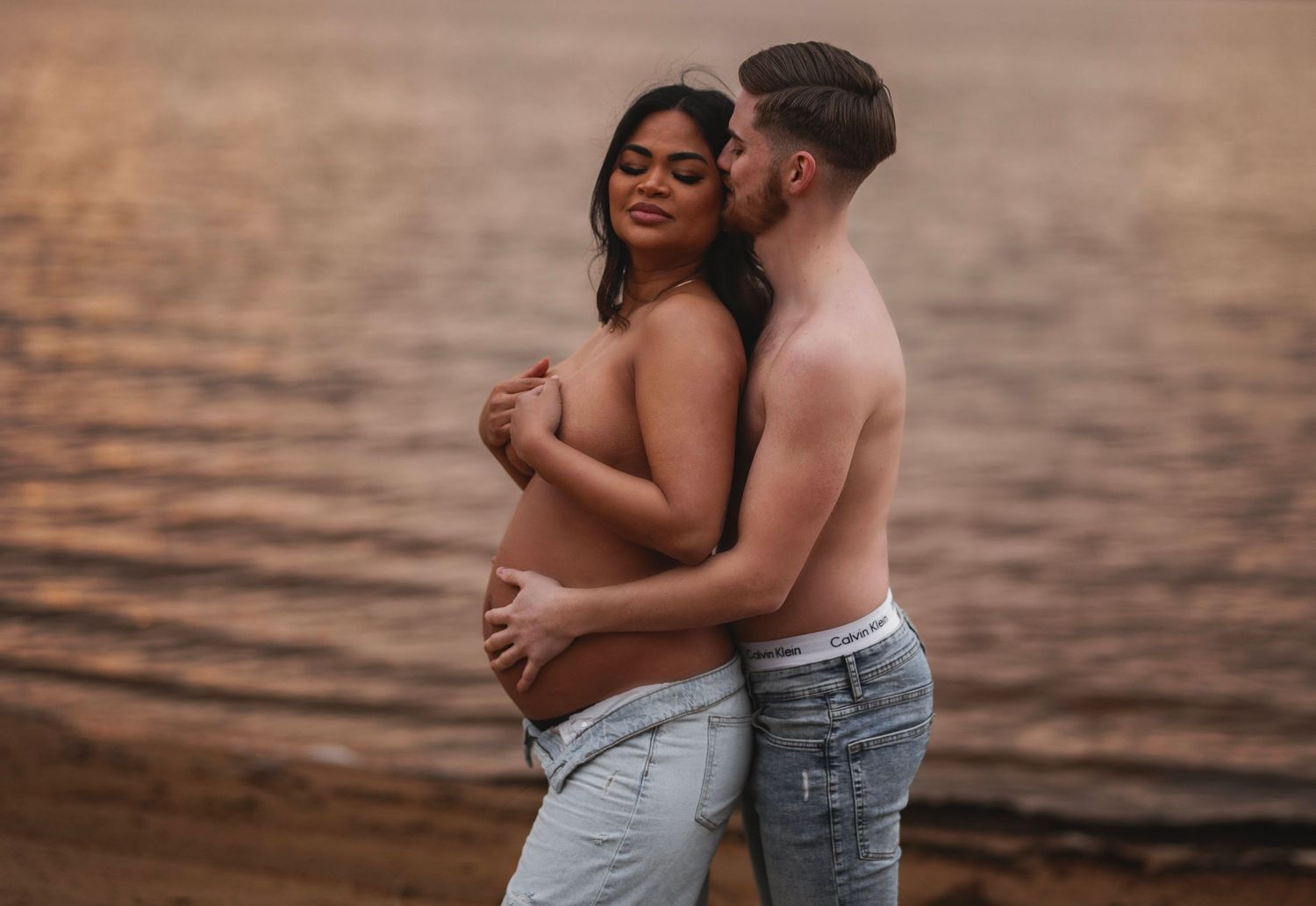 An intimate maternity photo of a couple embracing on a beach at golden hour.