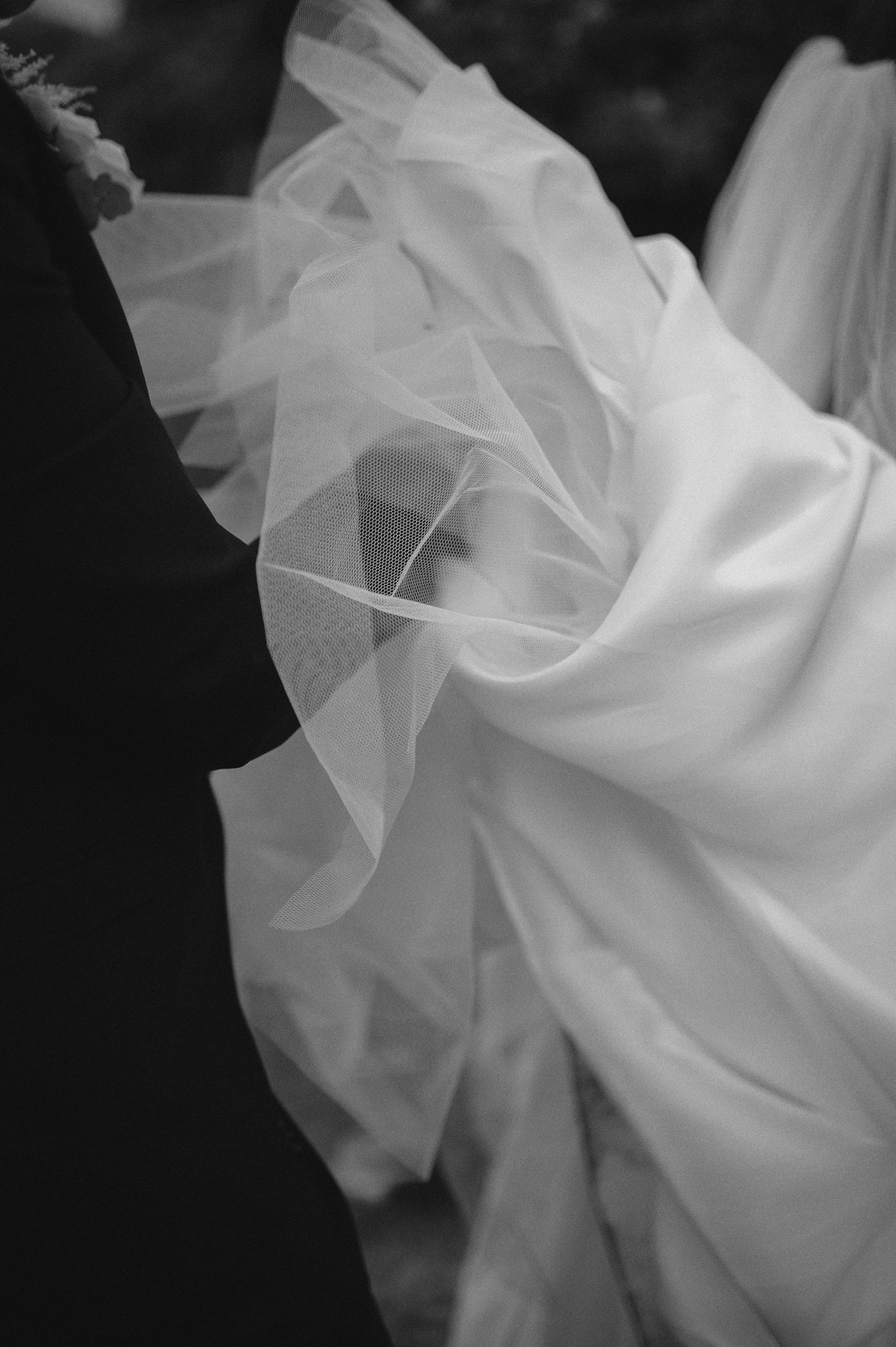 Black and white close-up of a flowing wedding dress veil billowing gracefully beside a groom's dark suit.