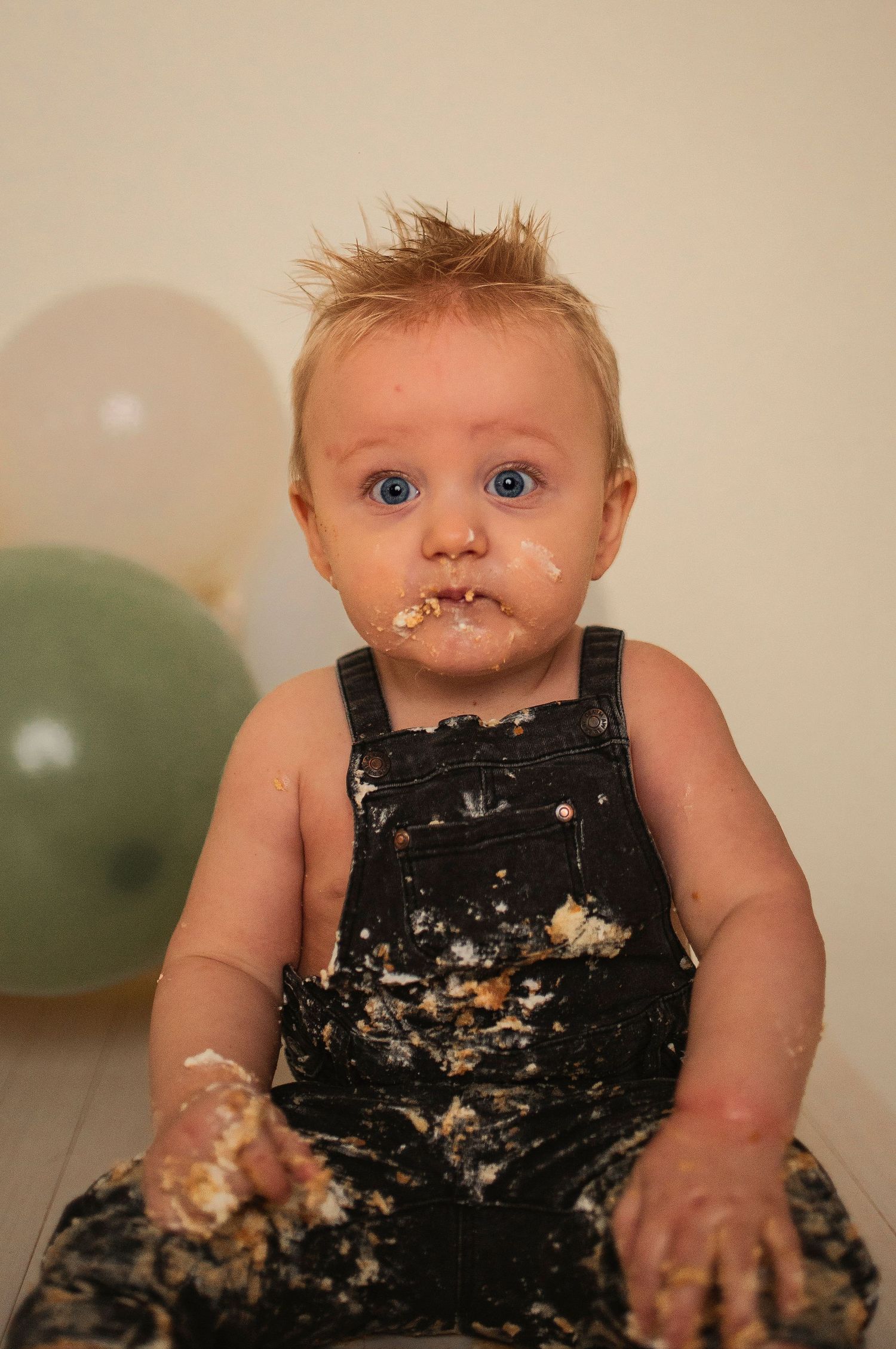 A messy cake smash photoshoot features a small child in black overalls covered in white frosting.