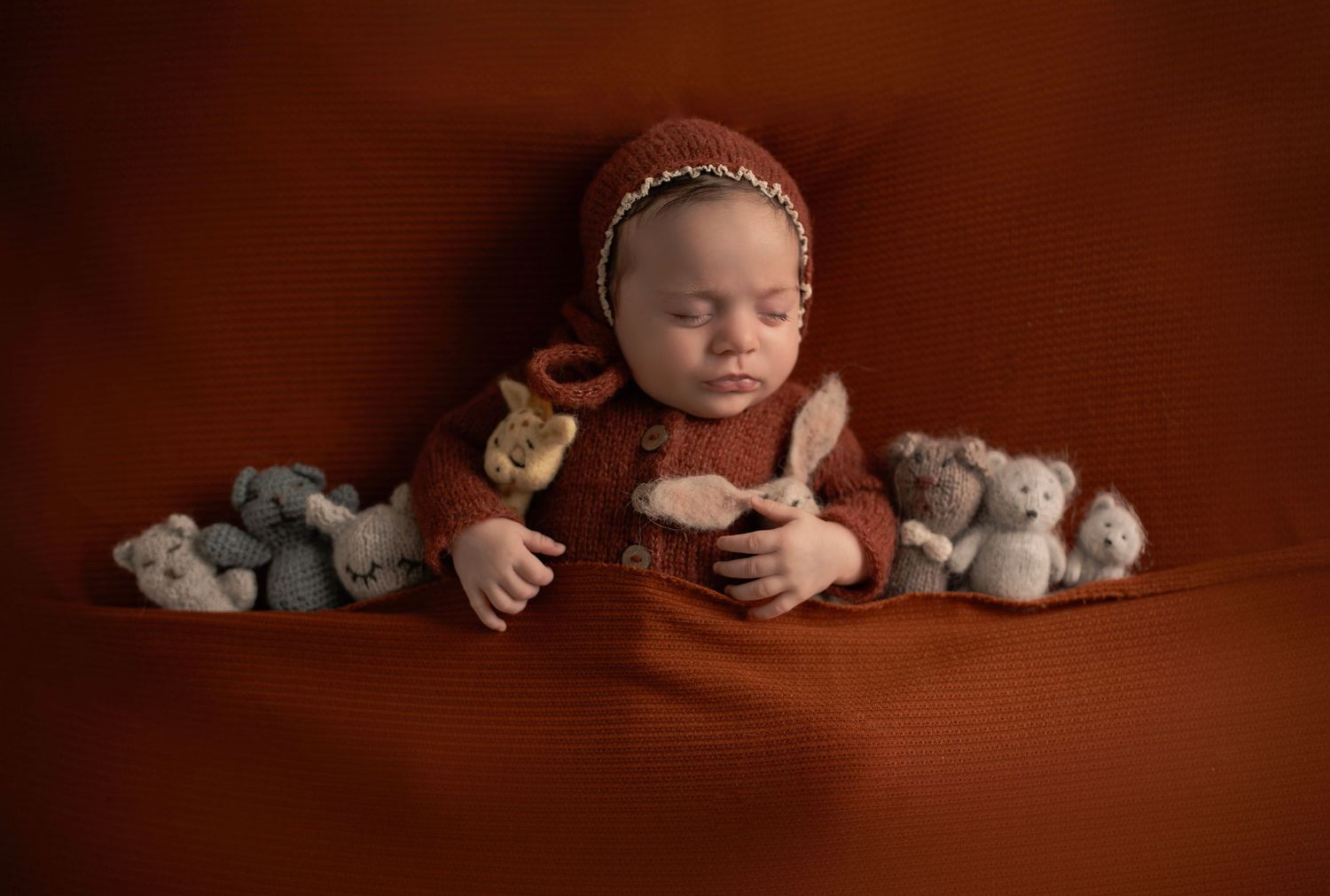 Infant in red sweater surrounded by soft plush toys on brown backdrop.